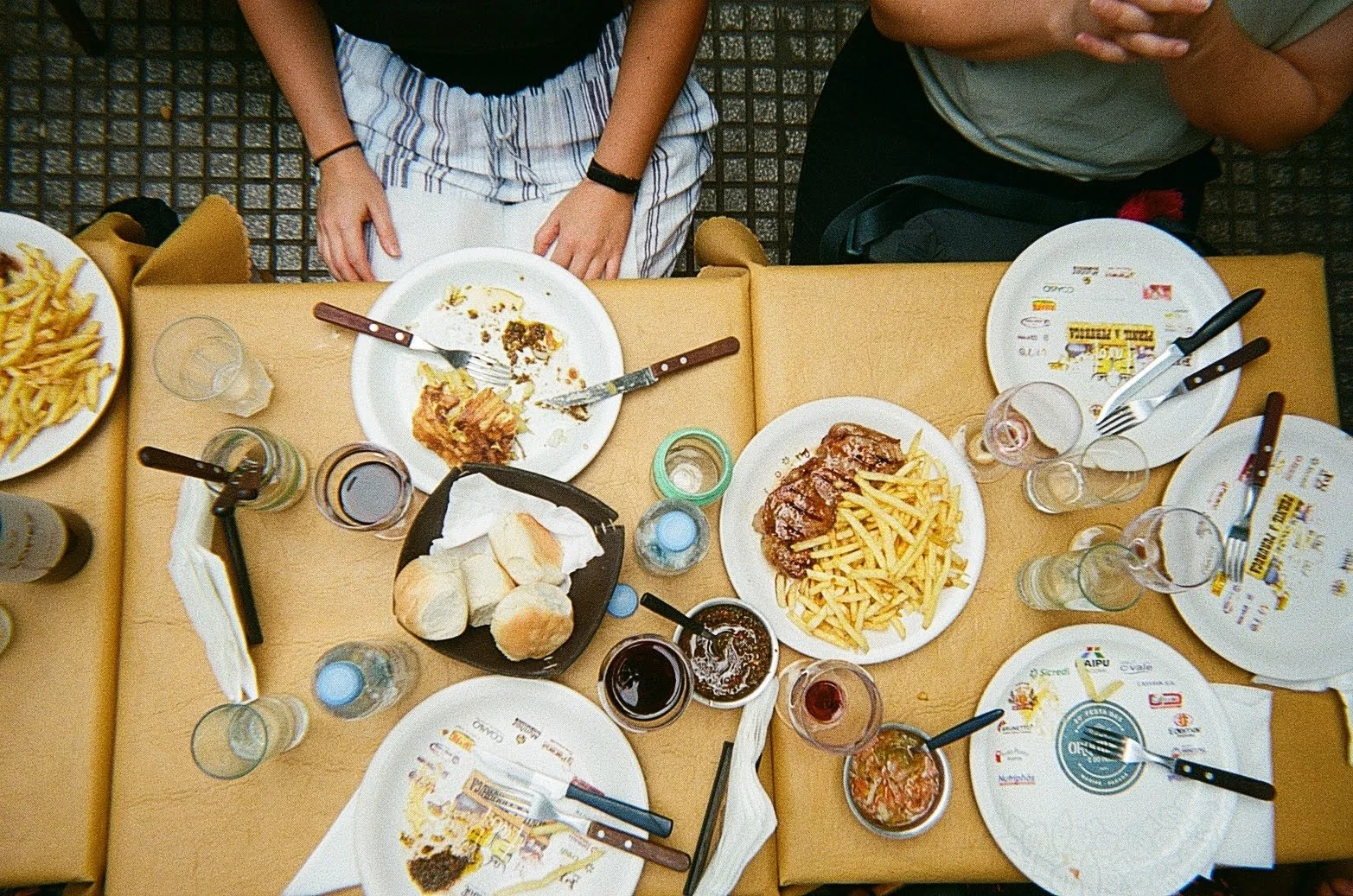 Top-down view of a dinner table with partly eaten food including fries, a steak with fries, bread rolls, and various beverages, with people sitting around the table.