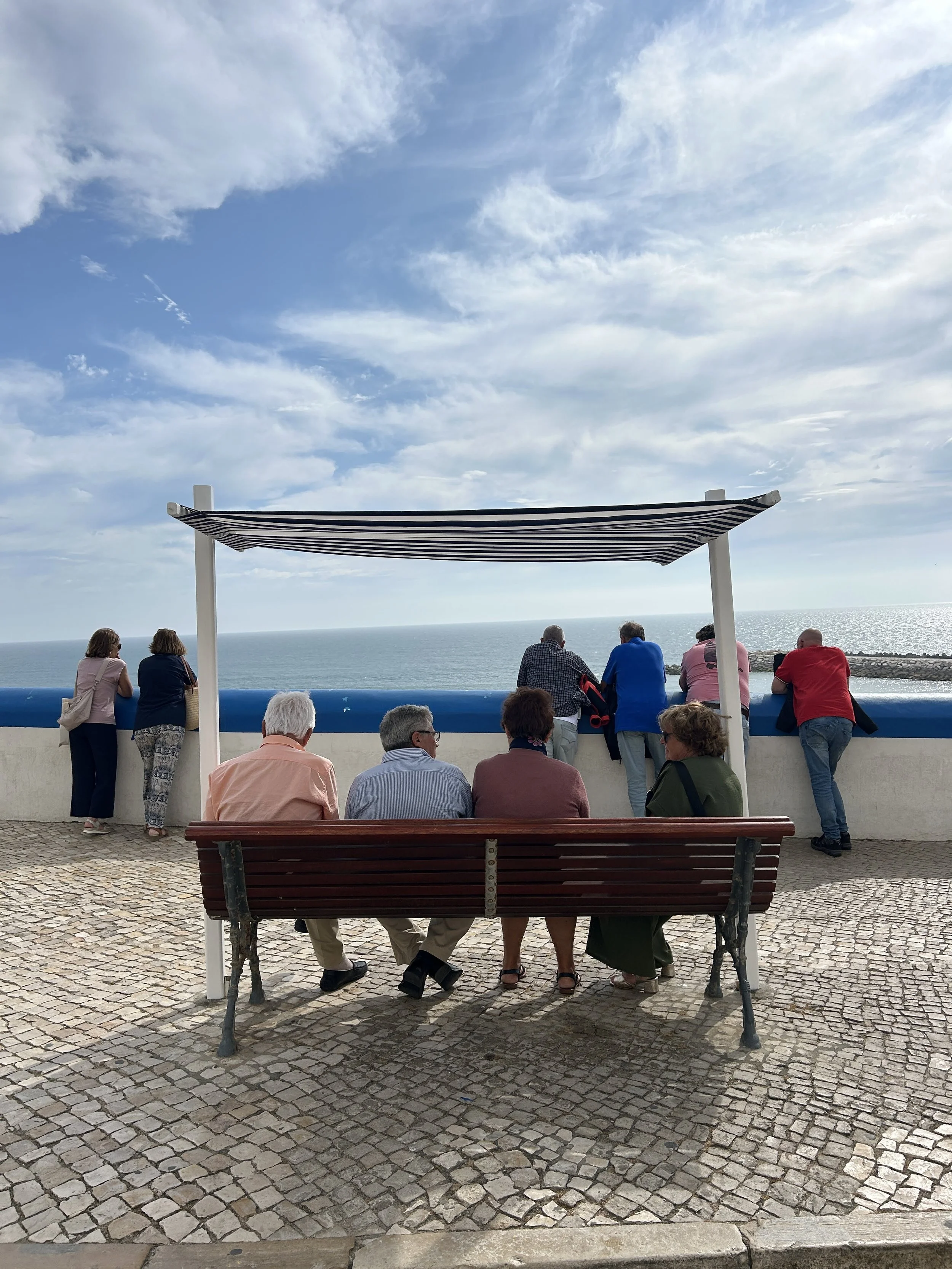 People sitting and standing by a waterfront on a cobblestone promenade, under a striped canopy with an ocean and cloudy sky in the background.