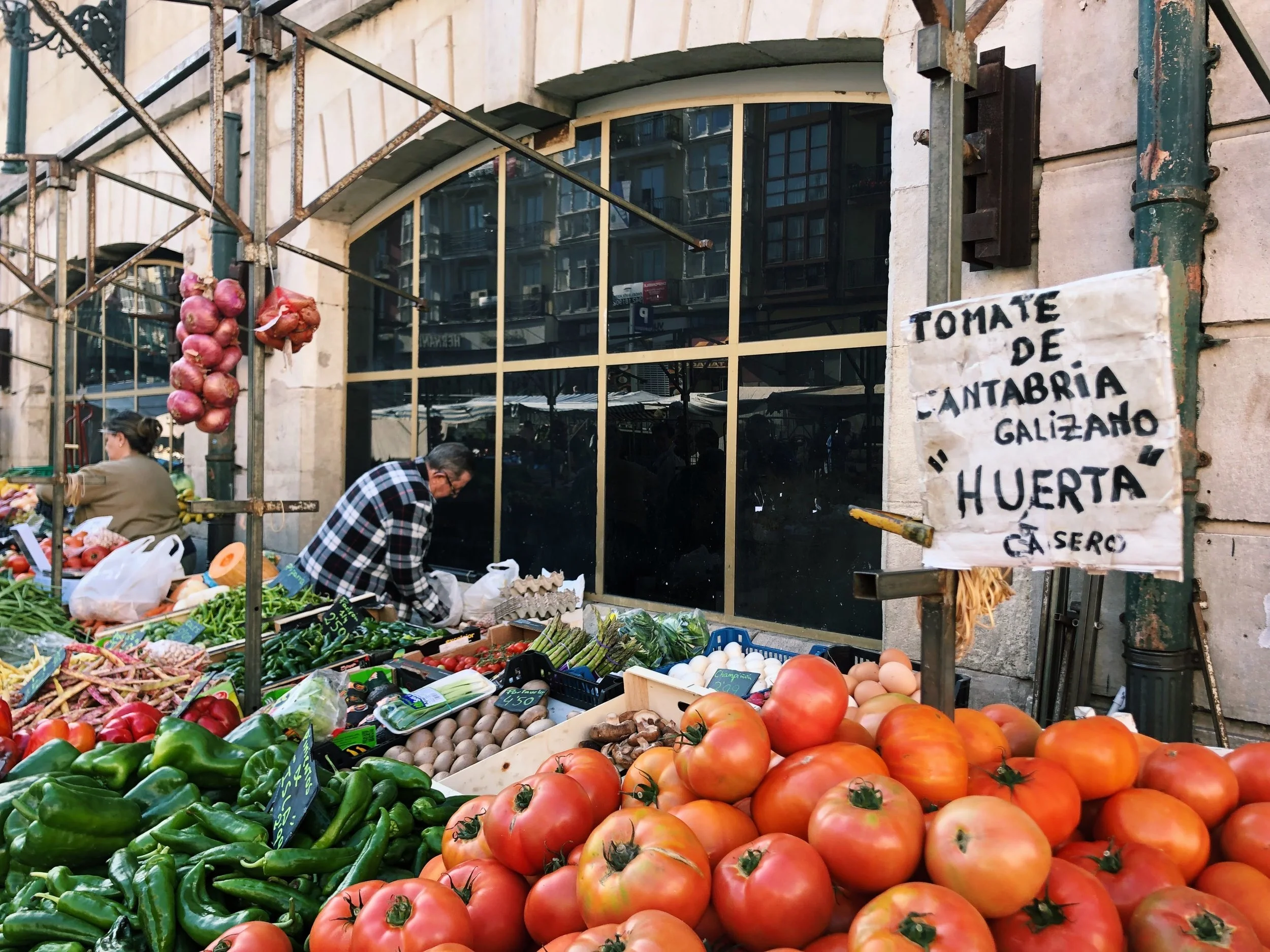 Market stall with fresh vegetables including tomatoes, peppers, cucumbers, and onions, with a woman shopping and a building reflected in the window behind.