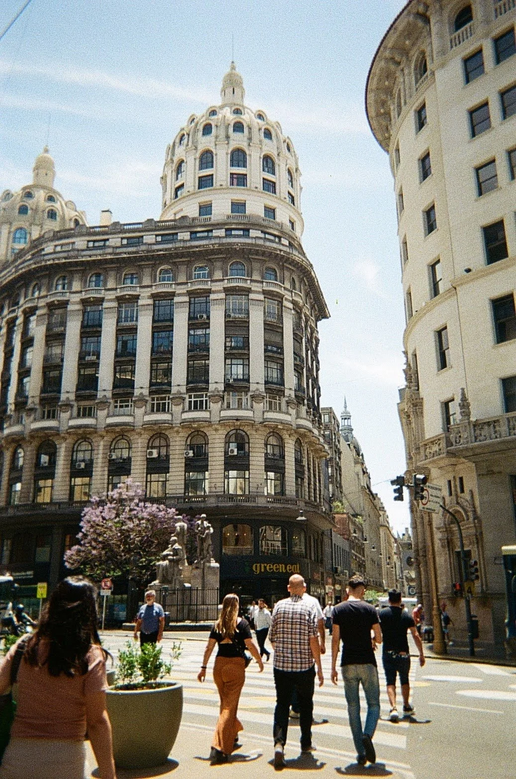 Downtown street scene with people crossing at crosswalk, large historic multi-story buildings, and clear sky.