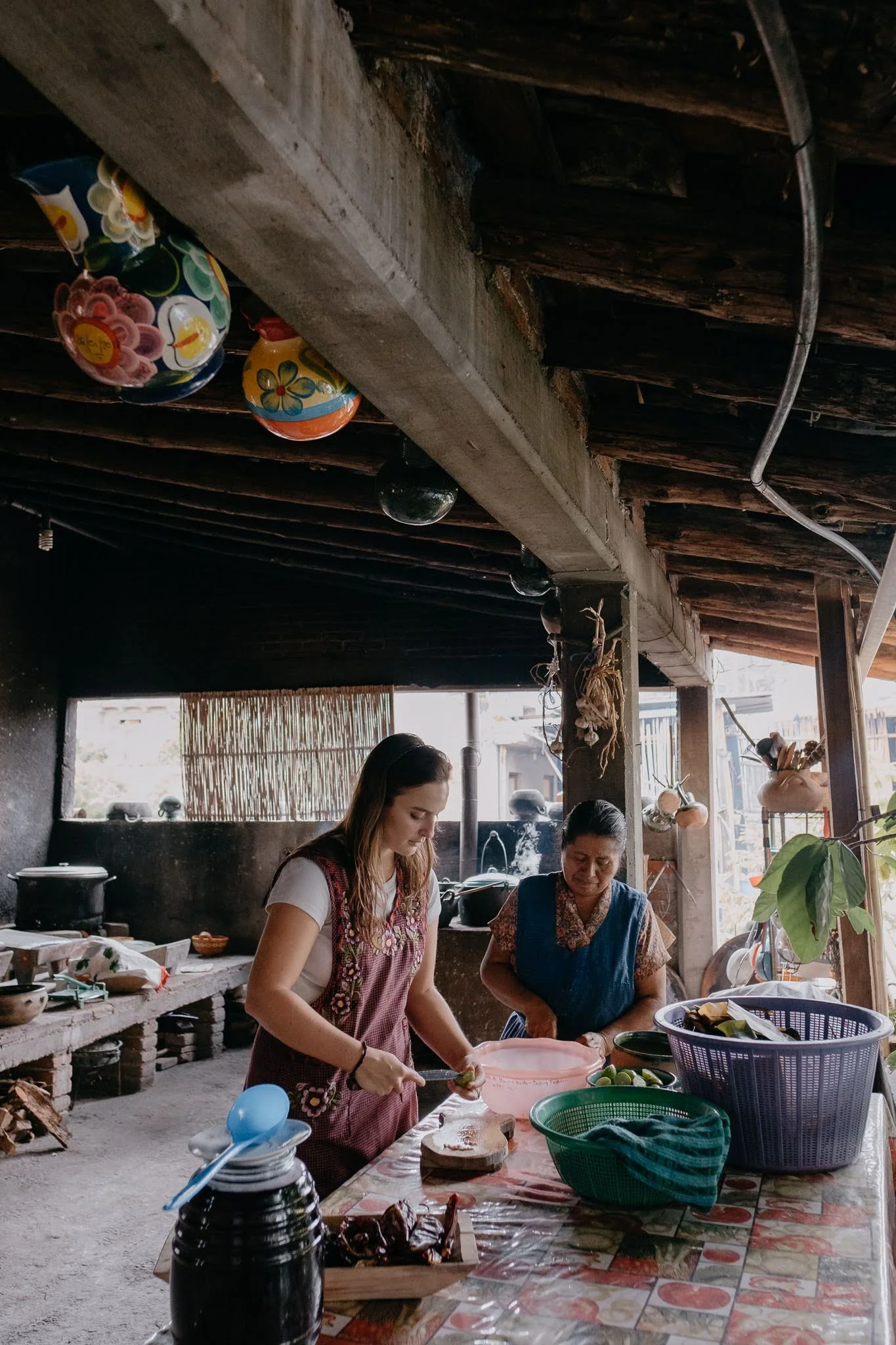 Two women preparing food at a table in a rustic open-air kitchen. The women are focused on their tasks, with various plates, bowls, and utensils on the table. The kitchen has a concrete counter, hanging pots, and a bamboo screen in the background.