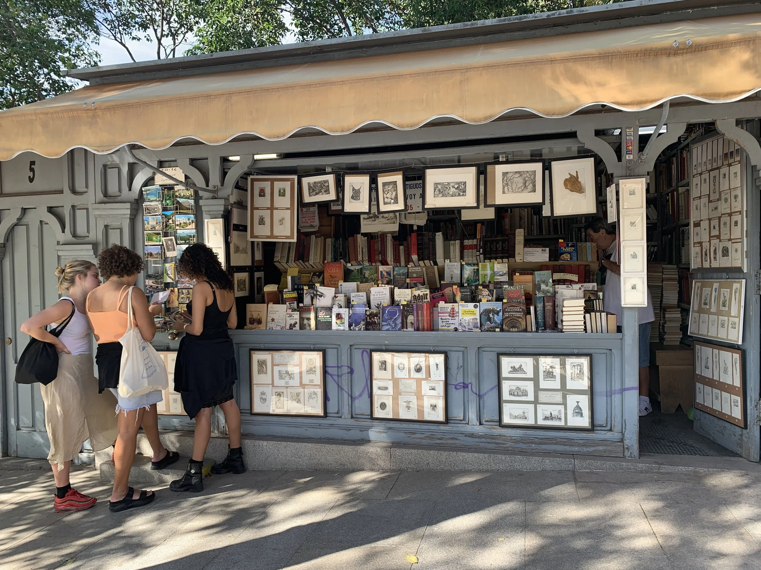 A small outdoor bookstore with a variety of books displayed on tables and framed artwork hanging above. Four women are browsing and chatting near the bookstore, with trees and a blue sky in the background.