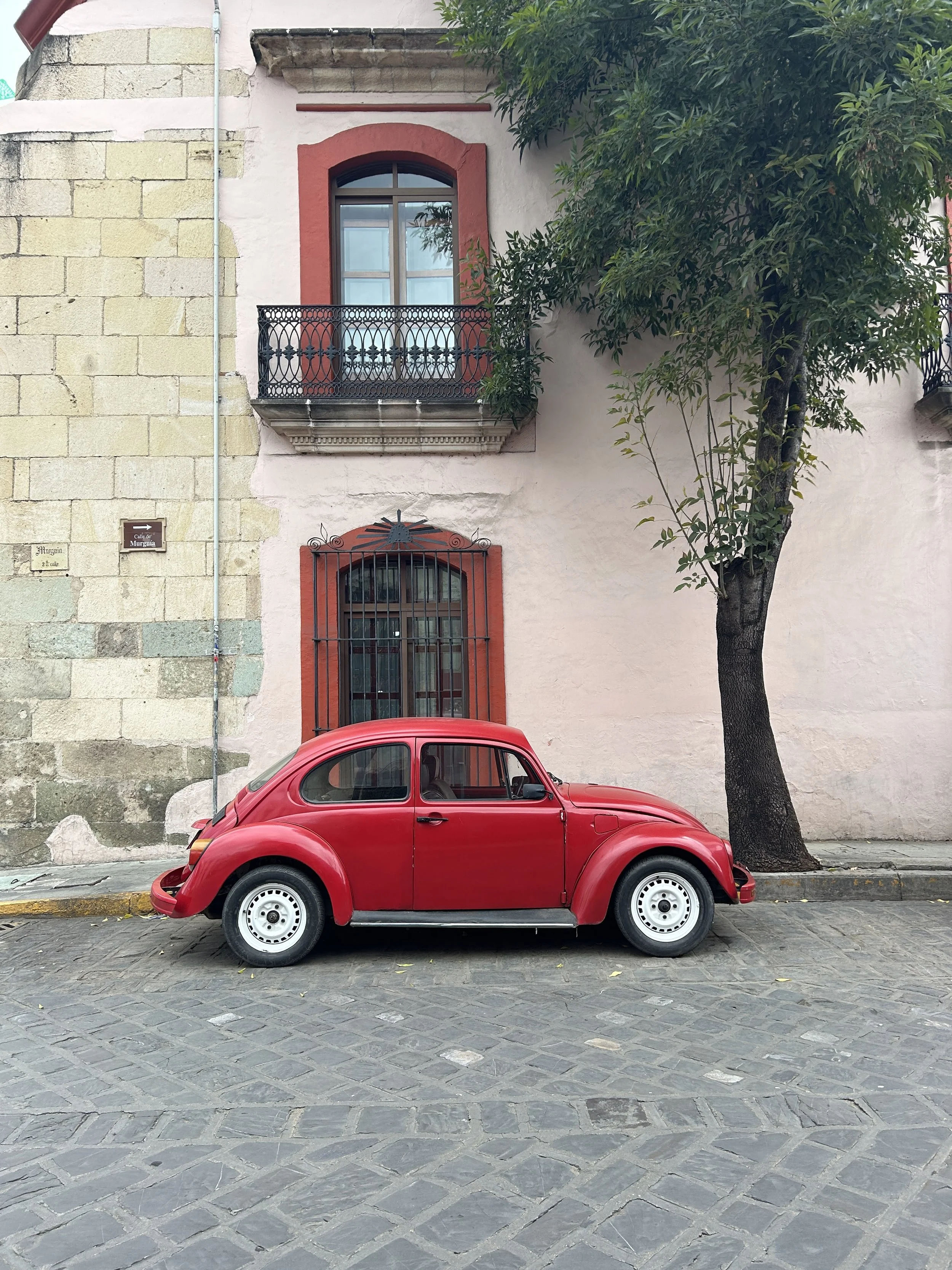 A red vintage Volkswagen Beetle parked on a cobblestone street in front of a building with a beige stone wall and light pink plaster. The building has two windows with black wrought iron balconies, and there is a large tree on the right side of the image.