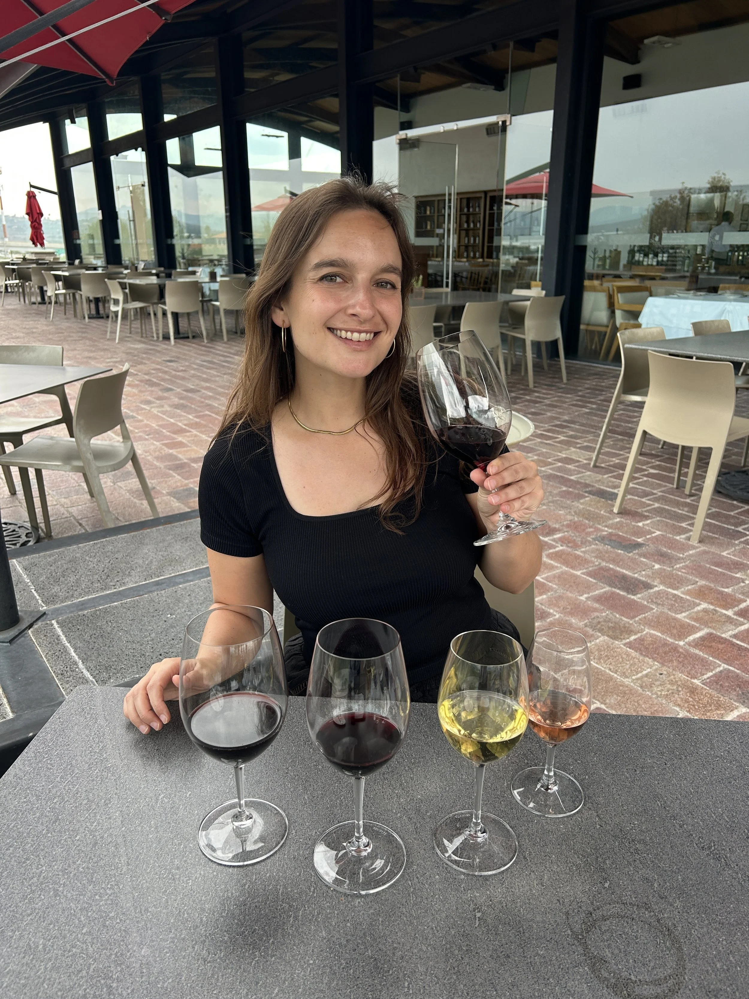 A smiling woman in a black top holding a glass of red wine, surrounded by four other glasses of red, white, and rosé wine on a table in an outdoor restaurant with brick flooring, large windows, and modern decor.