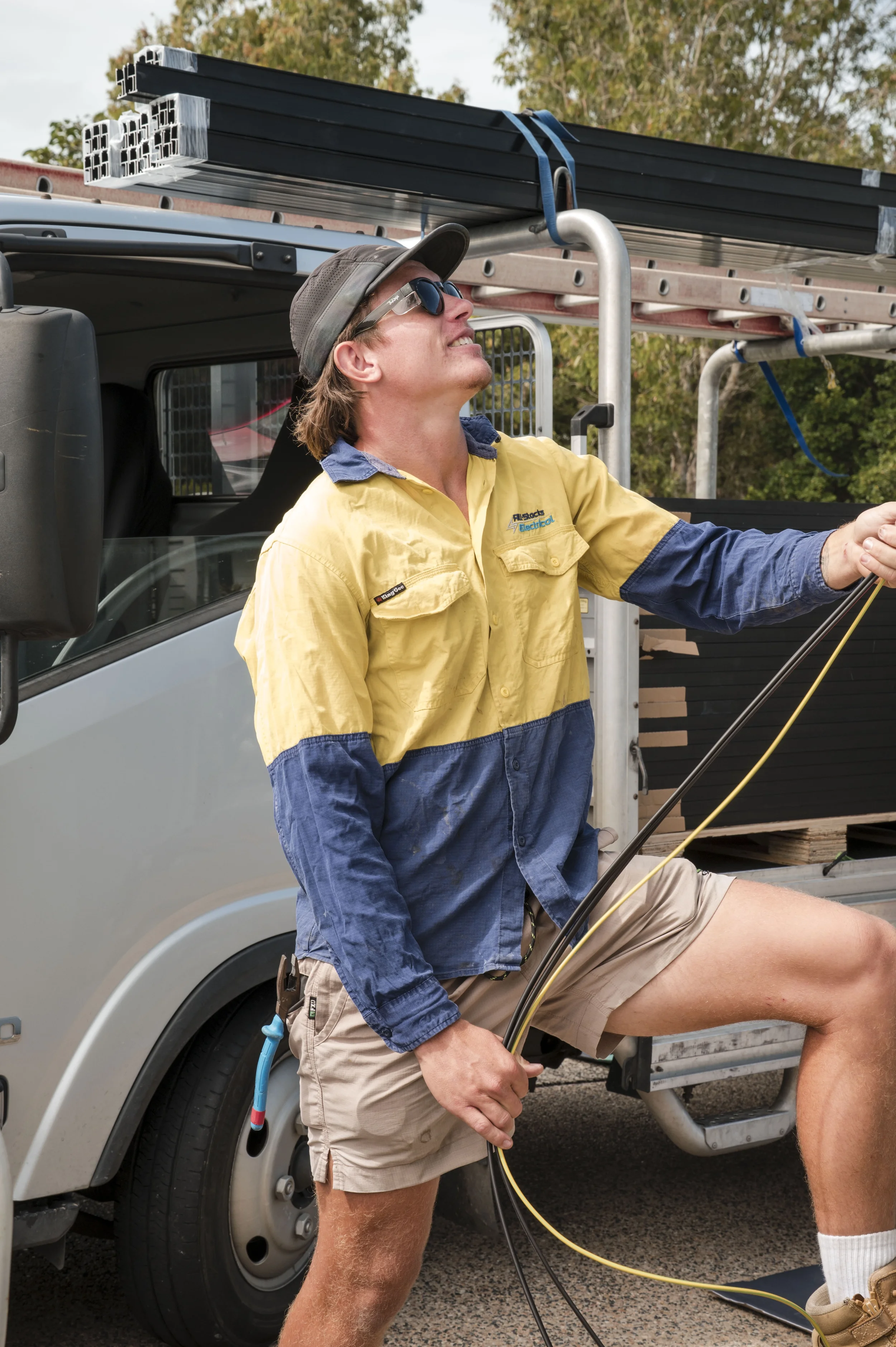 A man in beige shorts and a yellow work shirt with rolled-up sleeves, wearing a cap and sunglasses, standing. Worker is holding cables and appears to be working on electrical wiring.