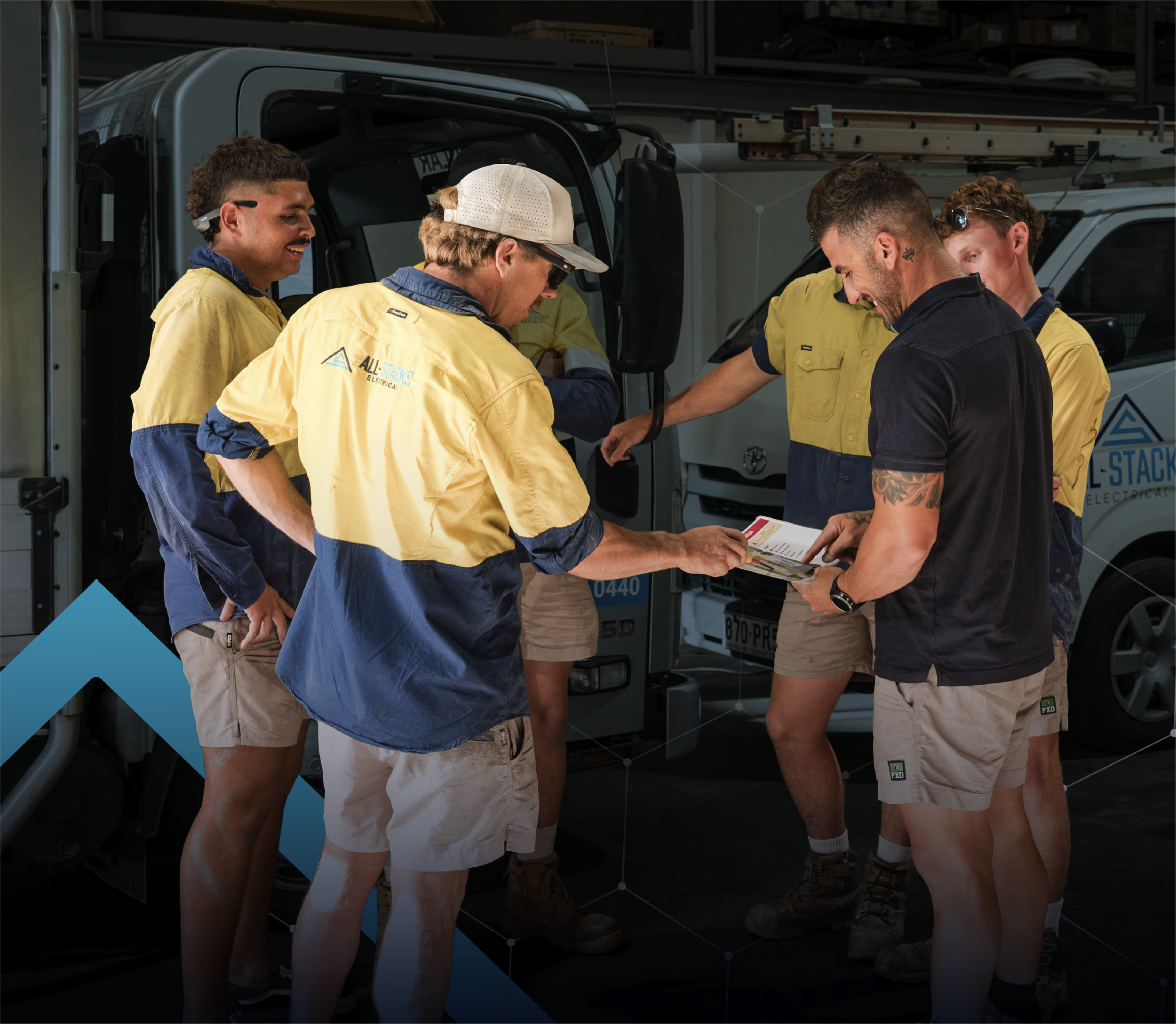 Group of five workers together in All Stacks Electrical workshop, wearing matching yellow and blue shirts, having a meeting and discussing jobs for the day, with vehicles and shelves in the background.