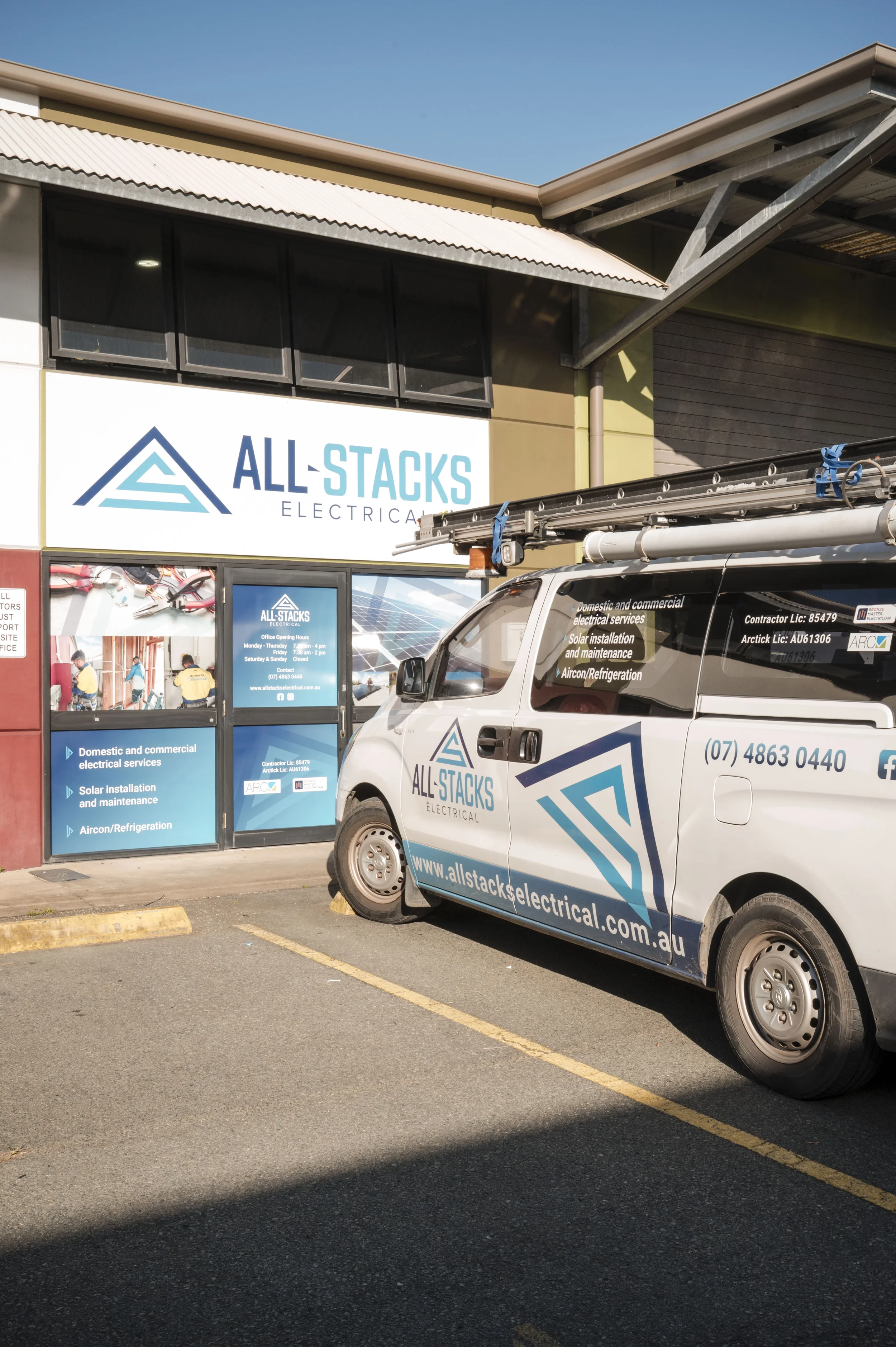 The image shows the storefront and parked vehicle of All-Stacks Electrical, an electrical service company. The building has a sign with the company logo and services listed, and a van with the company logo and website parked in front.