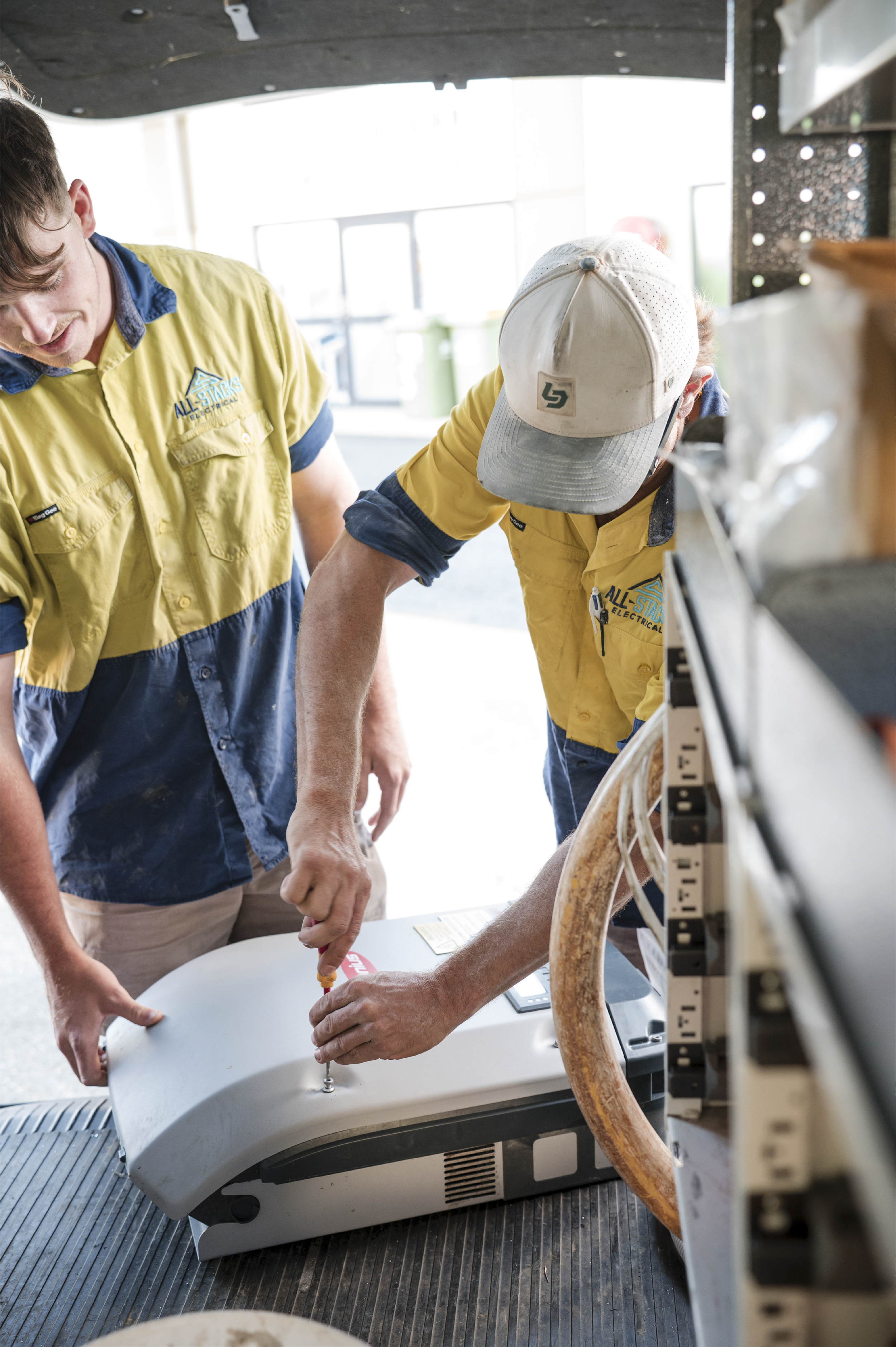 Two workers wearing yellow and blue uniforms are setting up an inverter, getting ready to install solar. Two works are fitting inverter inside a work vehicle with shelves and tools visible.