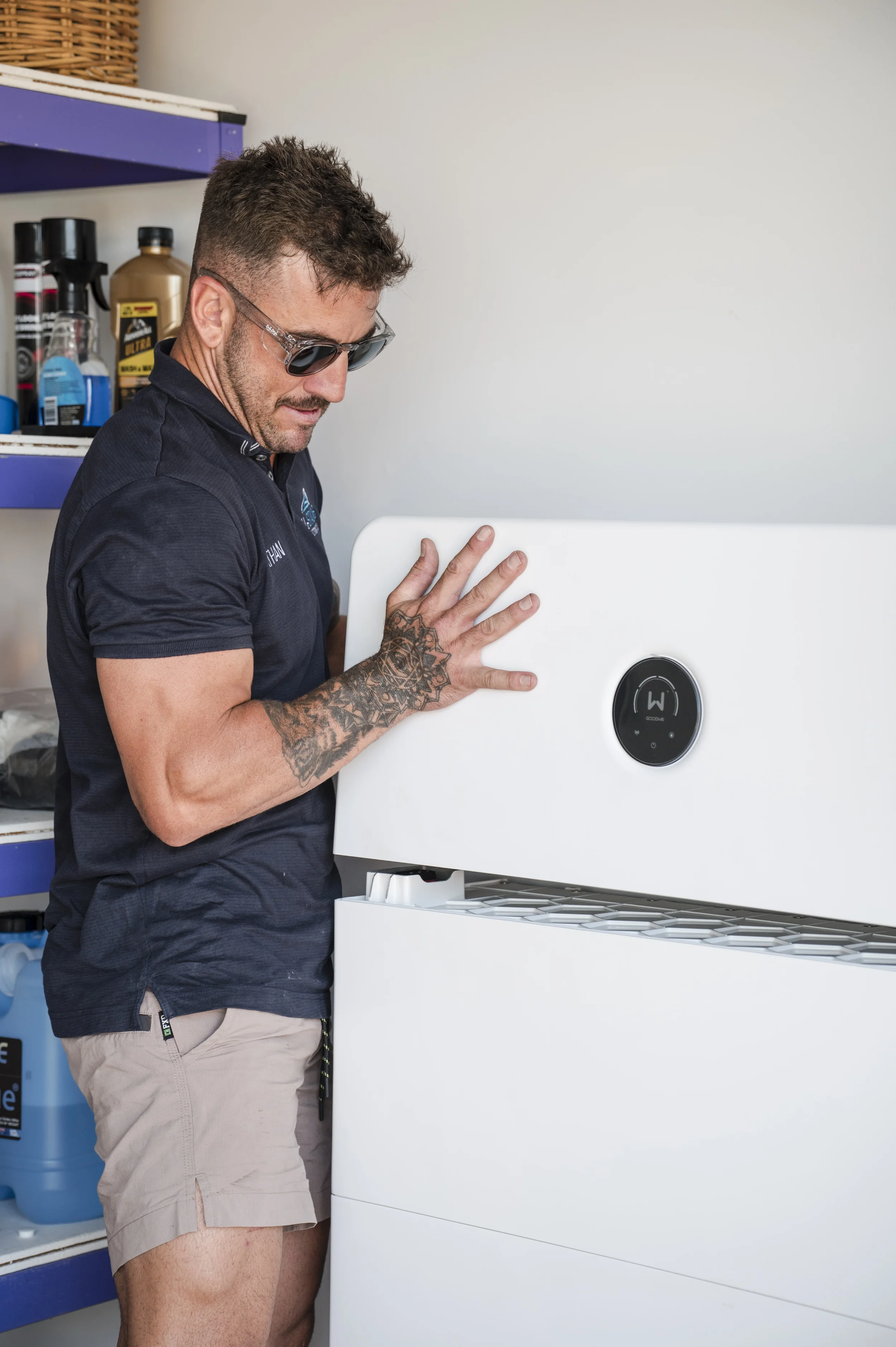 A man wearing sunglasses, a dark blue short-sleeved shirt, and beige shorts is installing a solar inverter inside a residential home