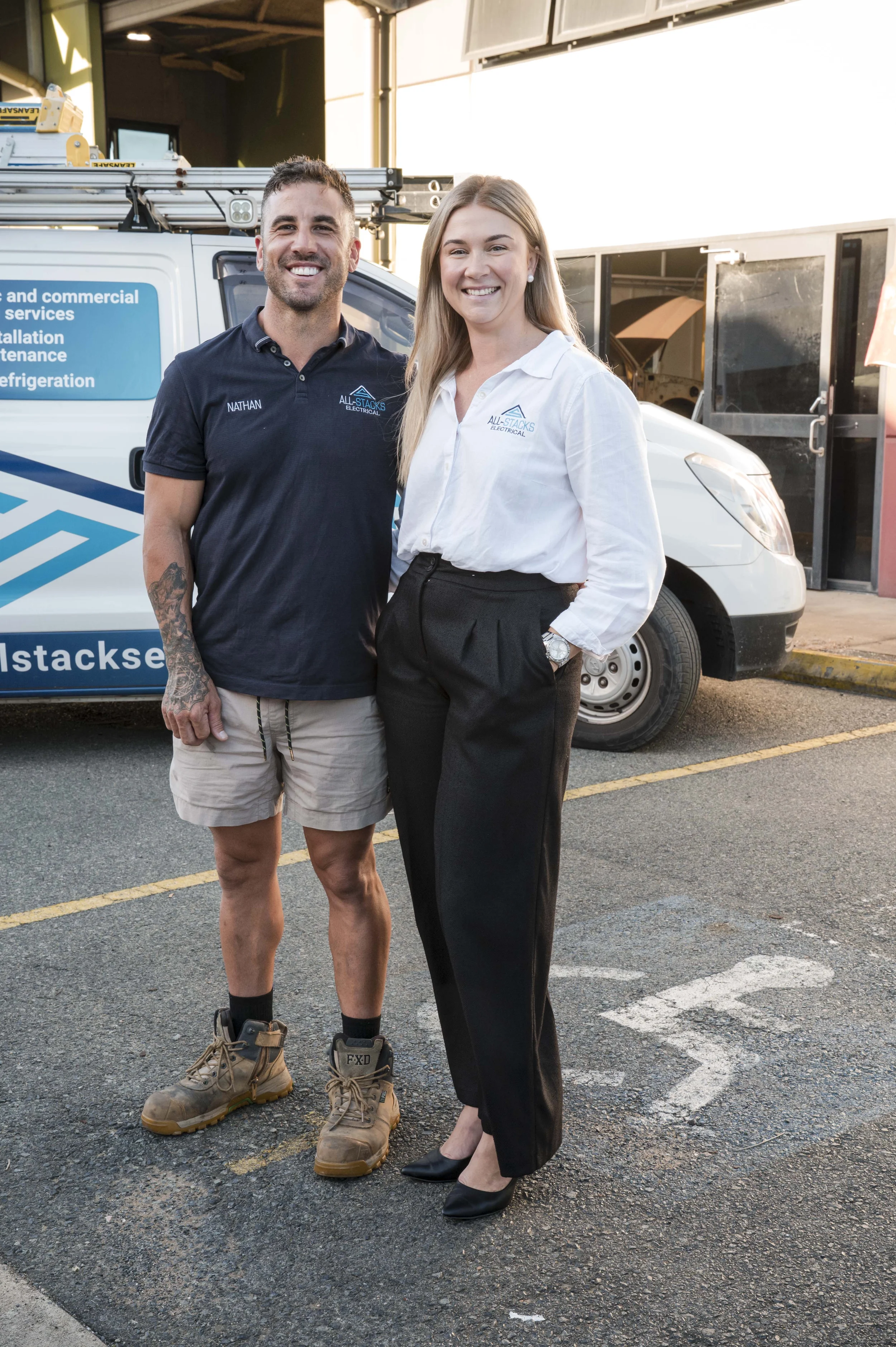 Two smiling professionals, a man in work attire and a woman in business clothing, stand in a parking lot with an electrical services van behind them, smiling at the camera.