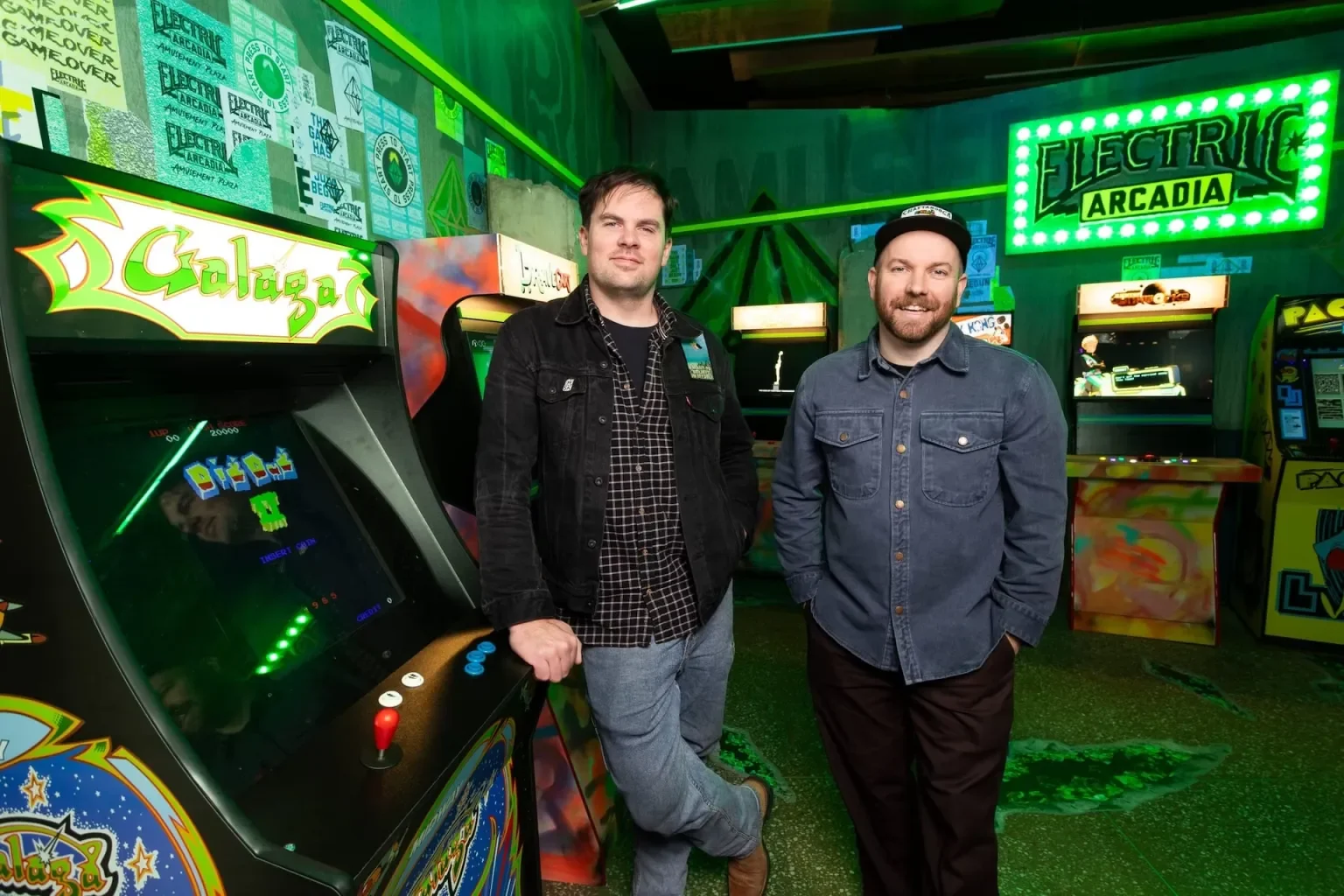Louie Roots and Callum Preston standing inside an arcade with retro arcade machines, neon green signs, and colorful lighting.