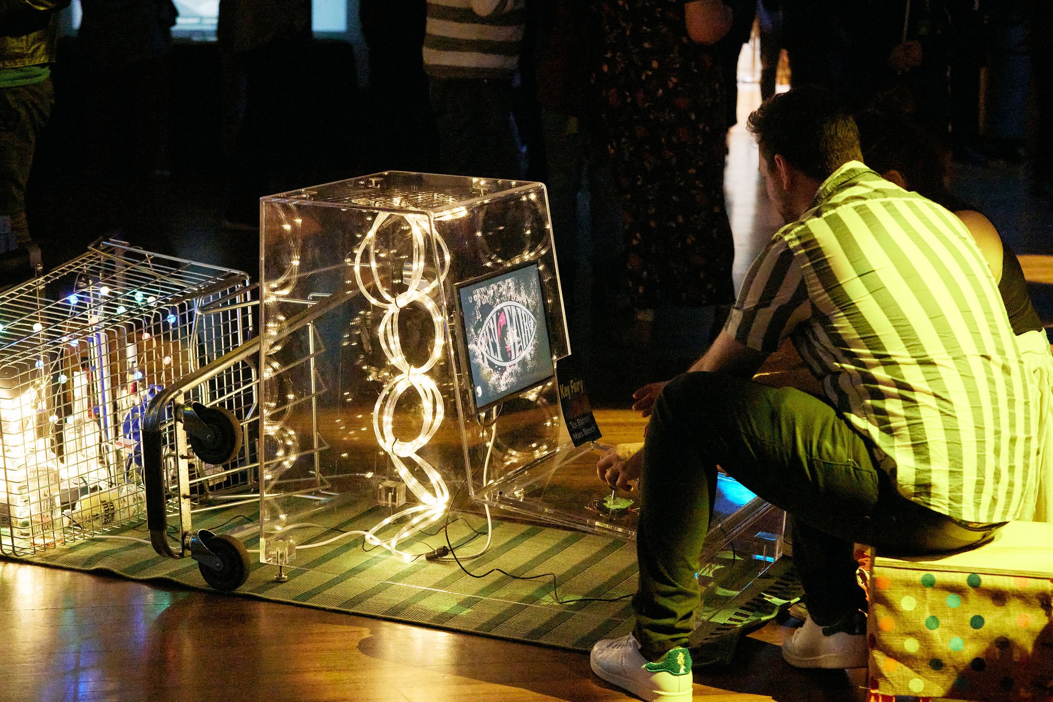 Acrylic Arcade Cabinet with custom LED internals and unique interactive playful art being displayed next to an upturned shopping trolley