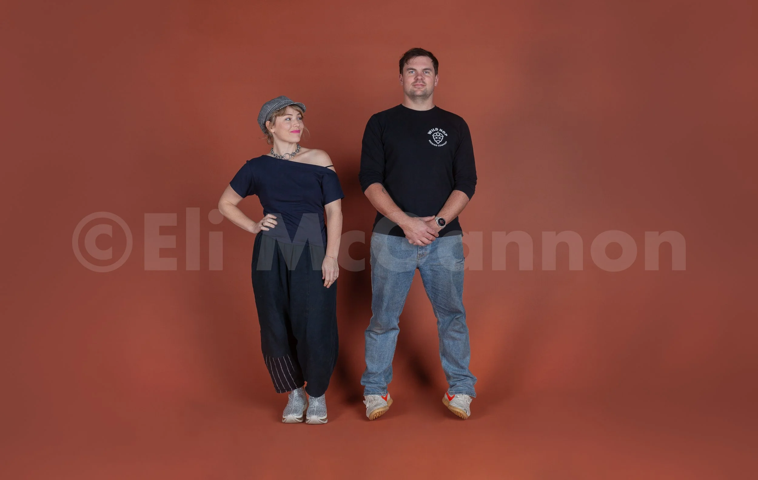 A woman and a man, Katie and Louie Roots, are standing side by side against a plain brown background.