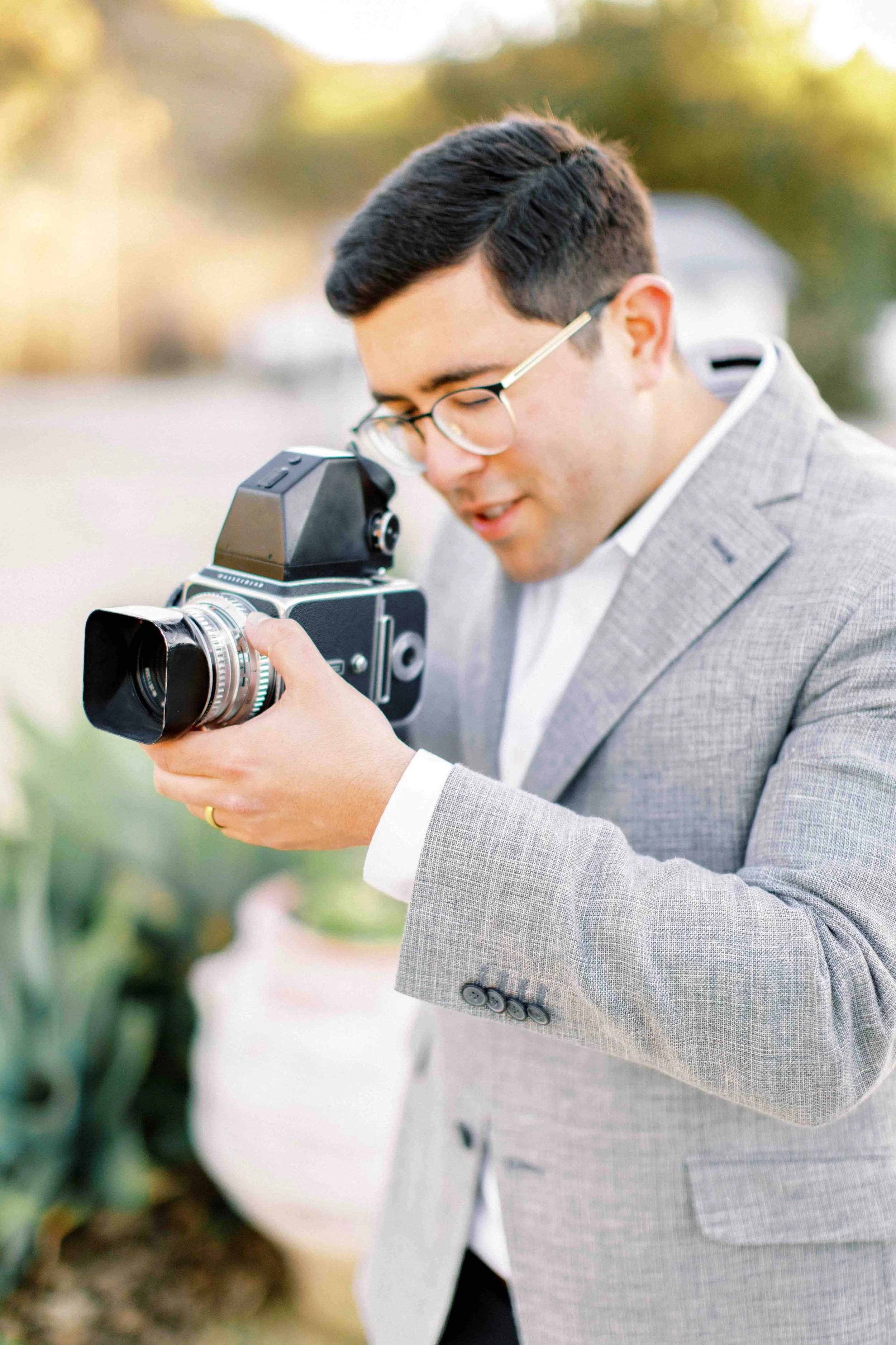 A man in a light gray checked suit jacket and glasses examines a vintage camera outdoors during daylight.