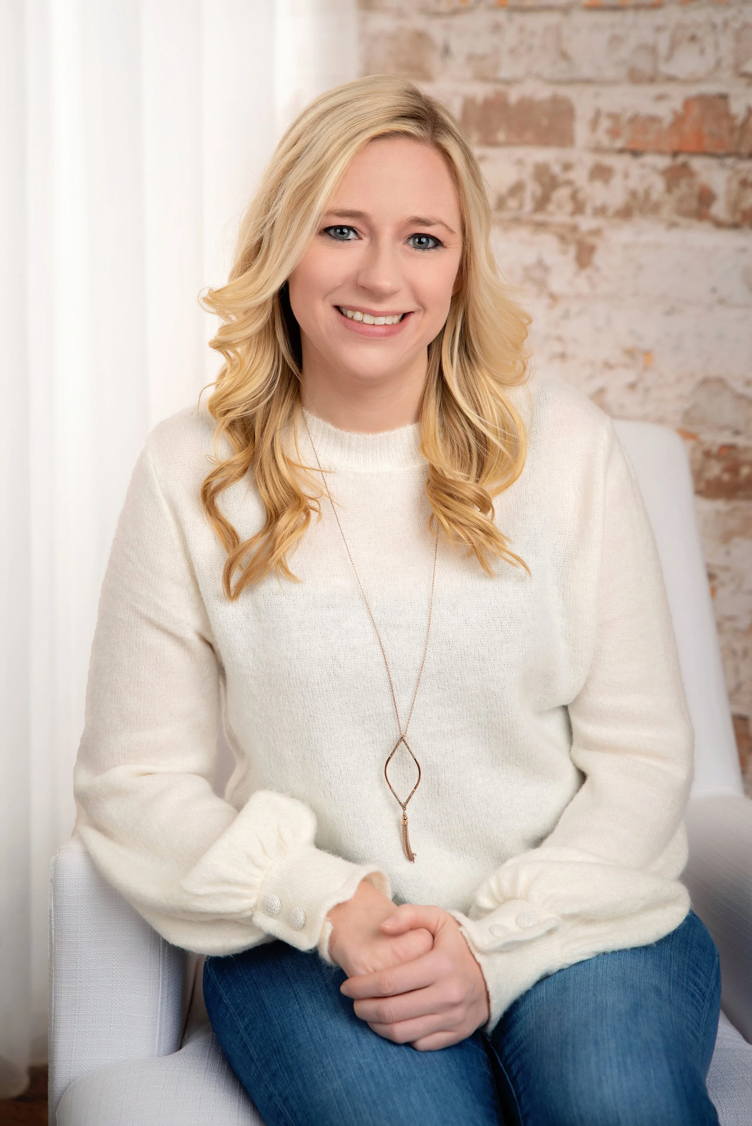A woman with blonde hair wearing a cream-colored sweater and blue jeans, sitting on a white chair, smiling, with a brick wall background.
