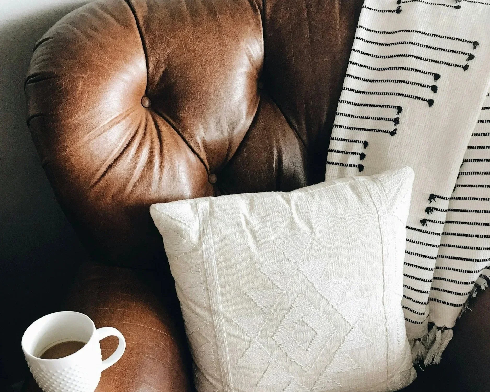 A cozy living room corner featuring a worn brown leather armchair, two white cups of coffee on a wooden table, a white textured pillow with a diamond pattern, and a curtain with black lines and tassels.
