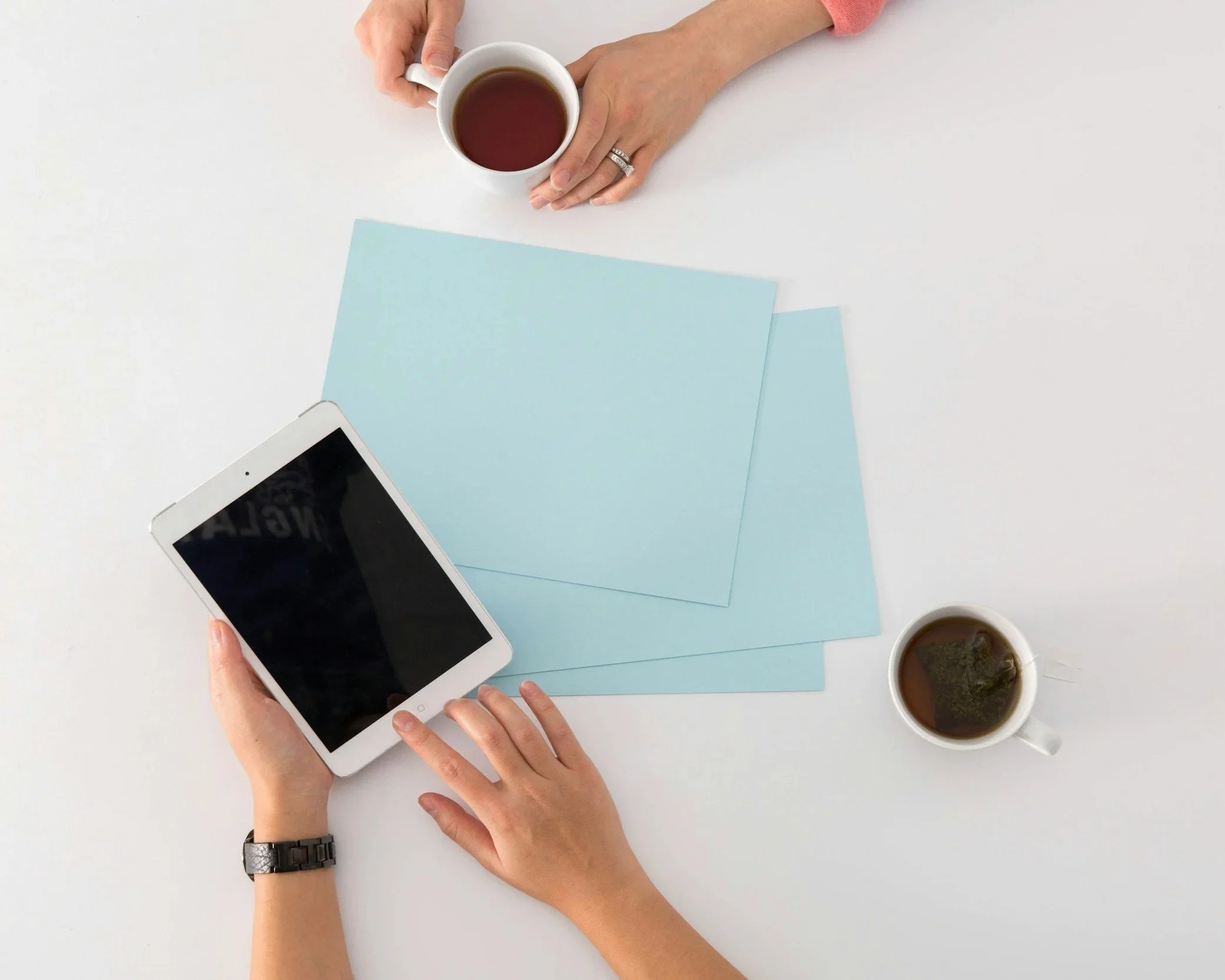 Top-down view of person holding smartphone with black screen, two cups of tea or coffee on white table, and light blue papers.