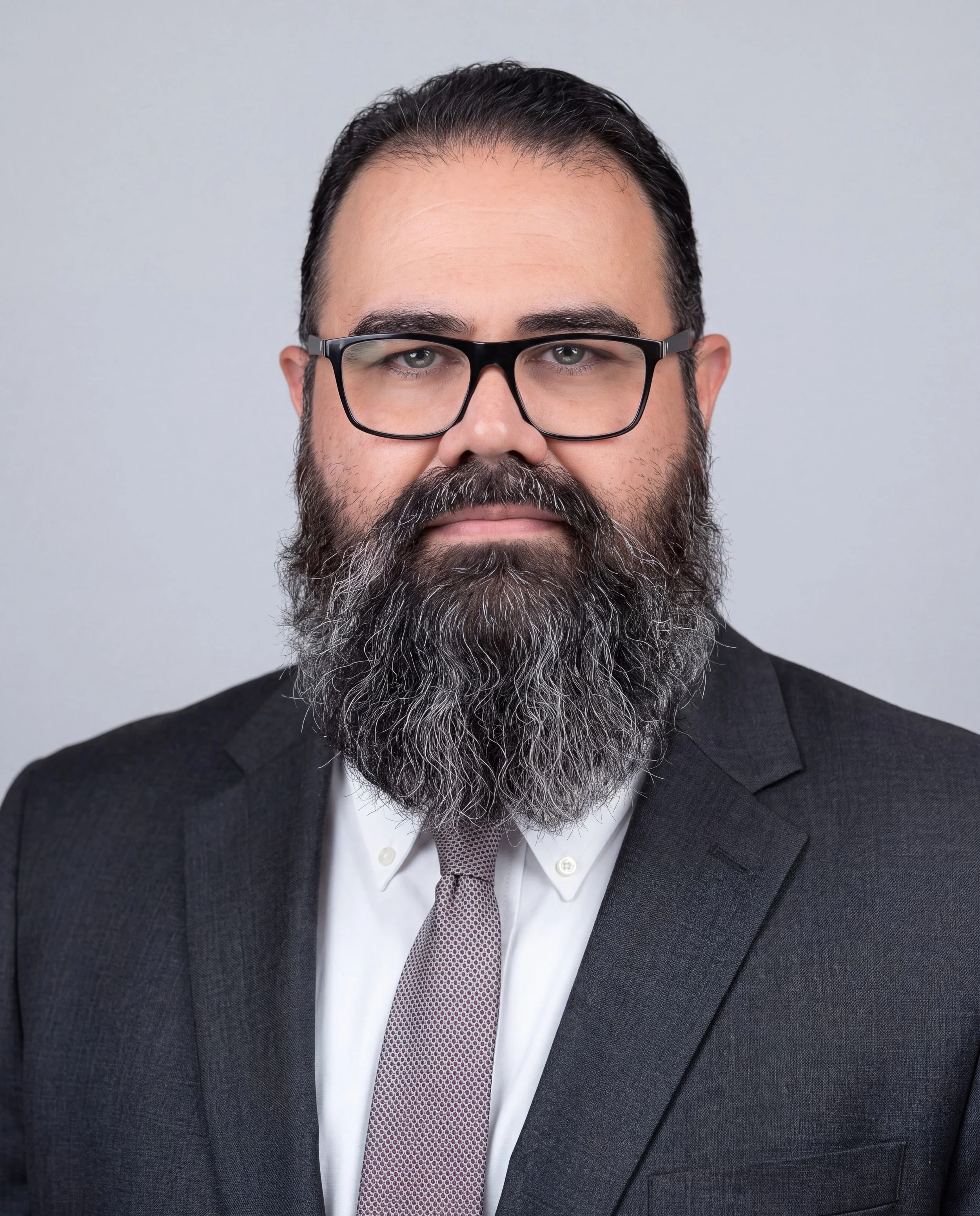 Close-up of a man with glasses, a beard, and a mustache, wearing a dark suit, white shirt, and a patterned tie, against a plain neutral background.