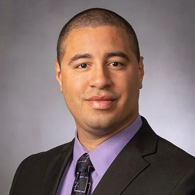Professional headshot of a man in a black suit, lavender shirt, and patterned tie, with short hair and a neutral background.