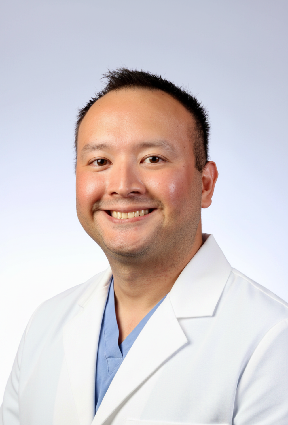 A smiling male healthcare professional in a white coat and blue scrubs against a light background.