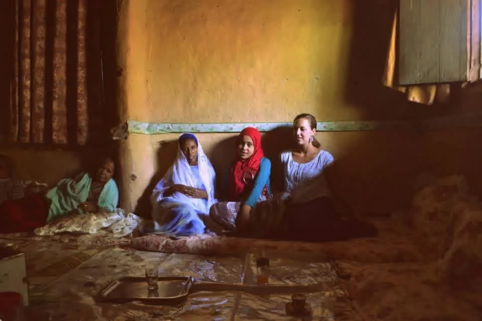 Four women sitting on a mattress inside a room with yellow walls, looking at the camera. The women are dressed in traditional clothing, and there are pillows and a tray on the mattress.