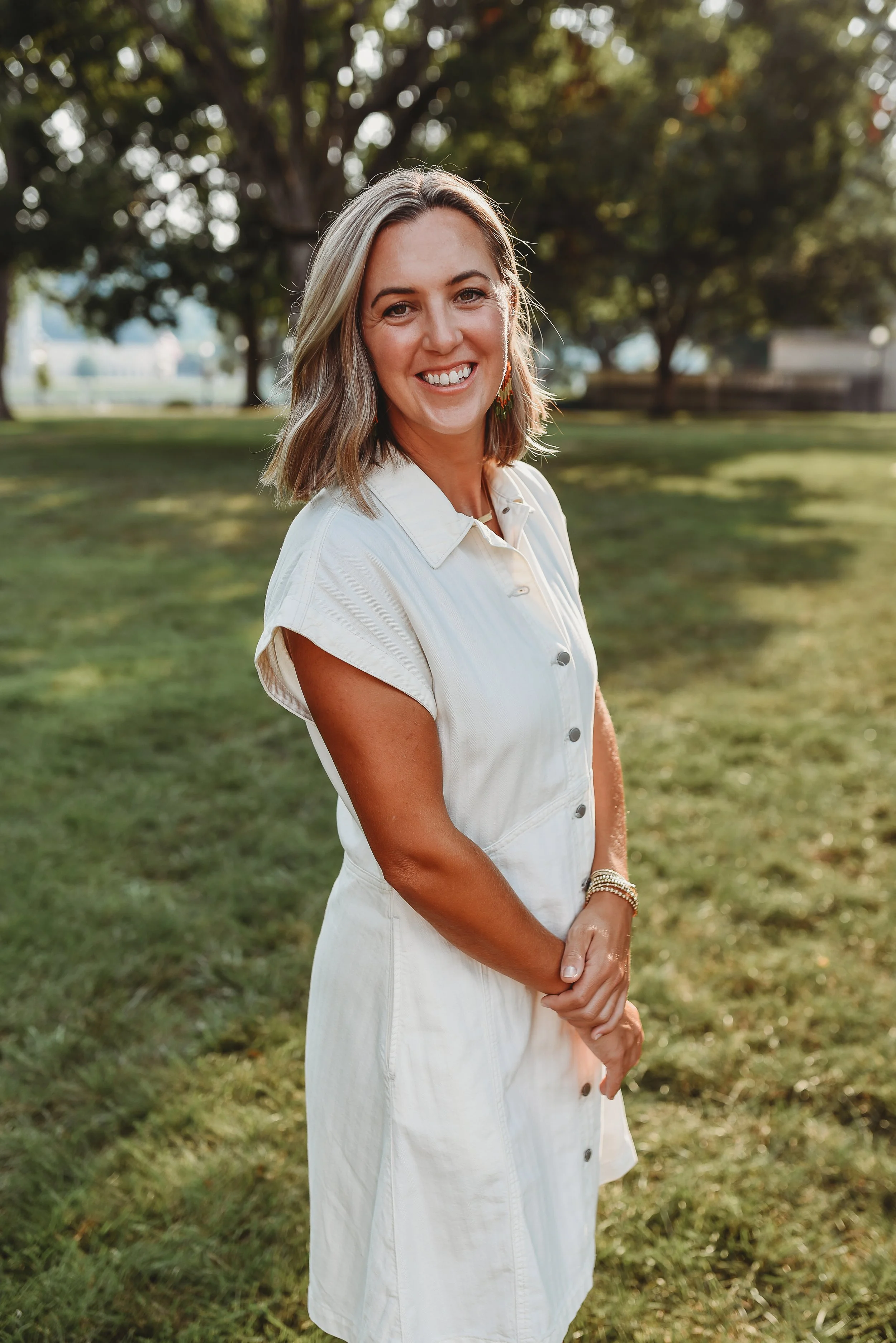 A woman with shoulder-length blonde hair, wearing a white dress with buttons, smiling outdoors with trees and grass in the background.