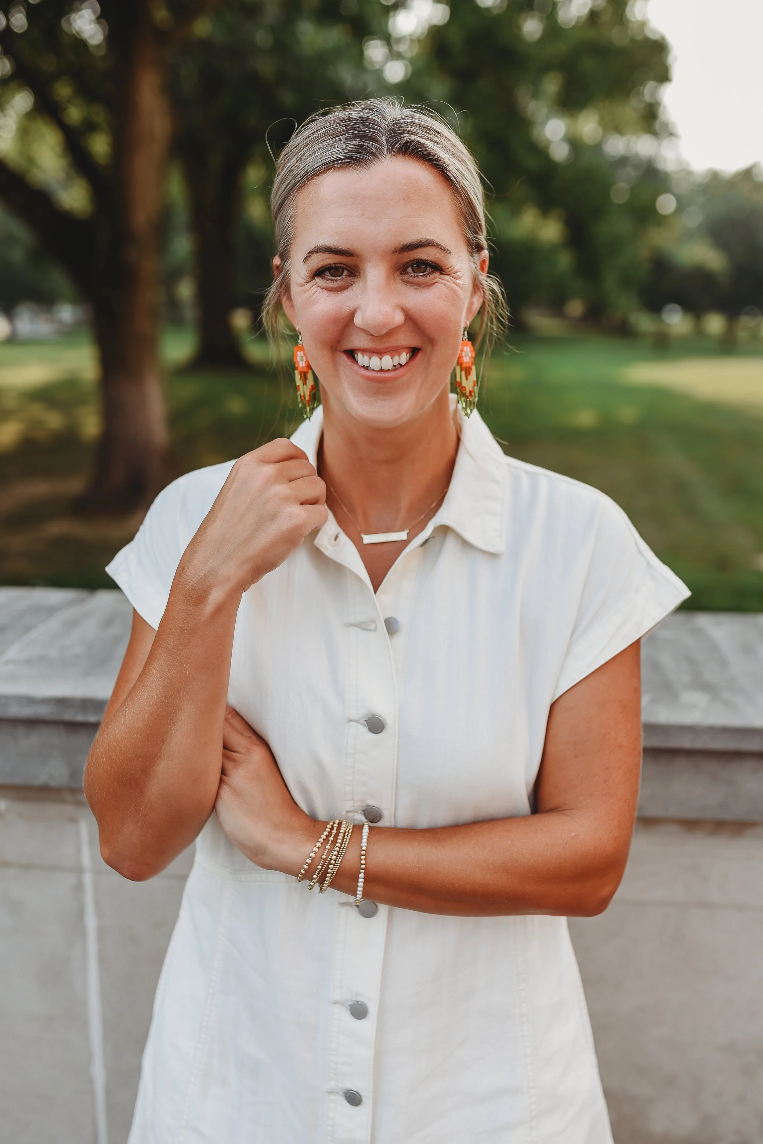 A woman with light brown hair, wearing a white button-up dress, colorful beaded earrings, and gold jewelry, smiling and standing outdoors with trees and grass in the background.