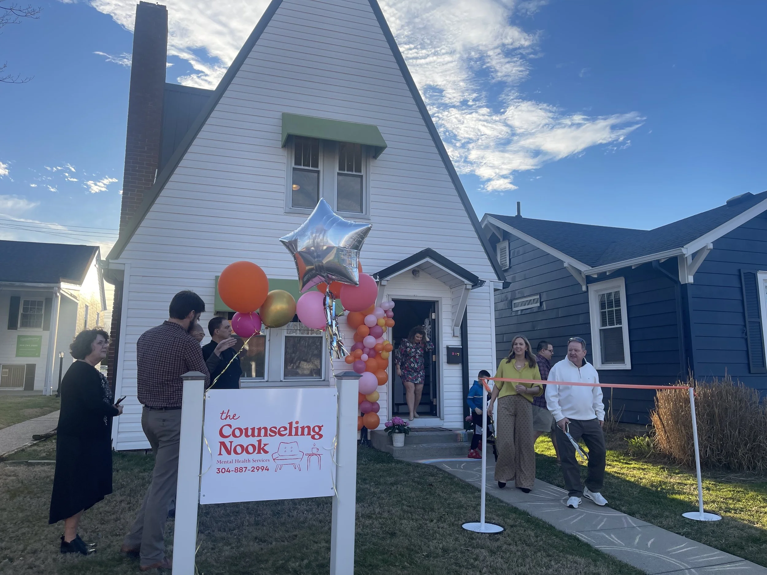 People gathered outside a white house decorated with pink, orange, and gold balloons for a ribbon-cutting event. There is a sign that reads 'the Counseling Nook' with a contact phone number below.