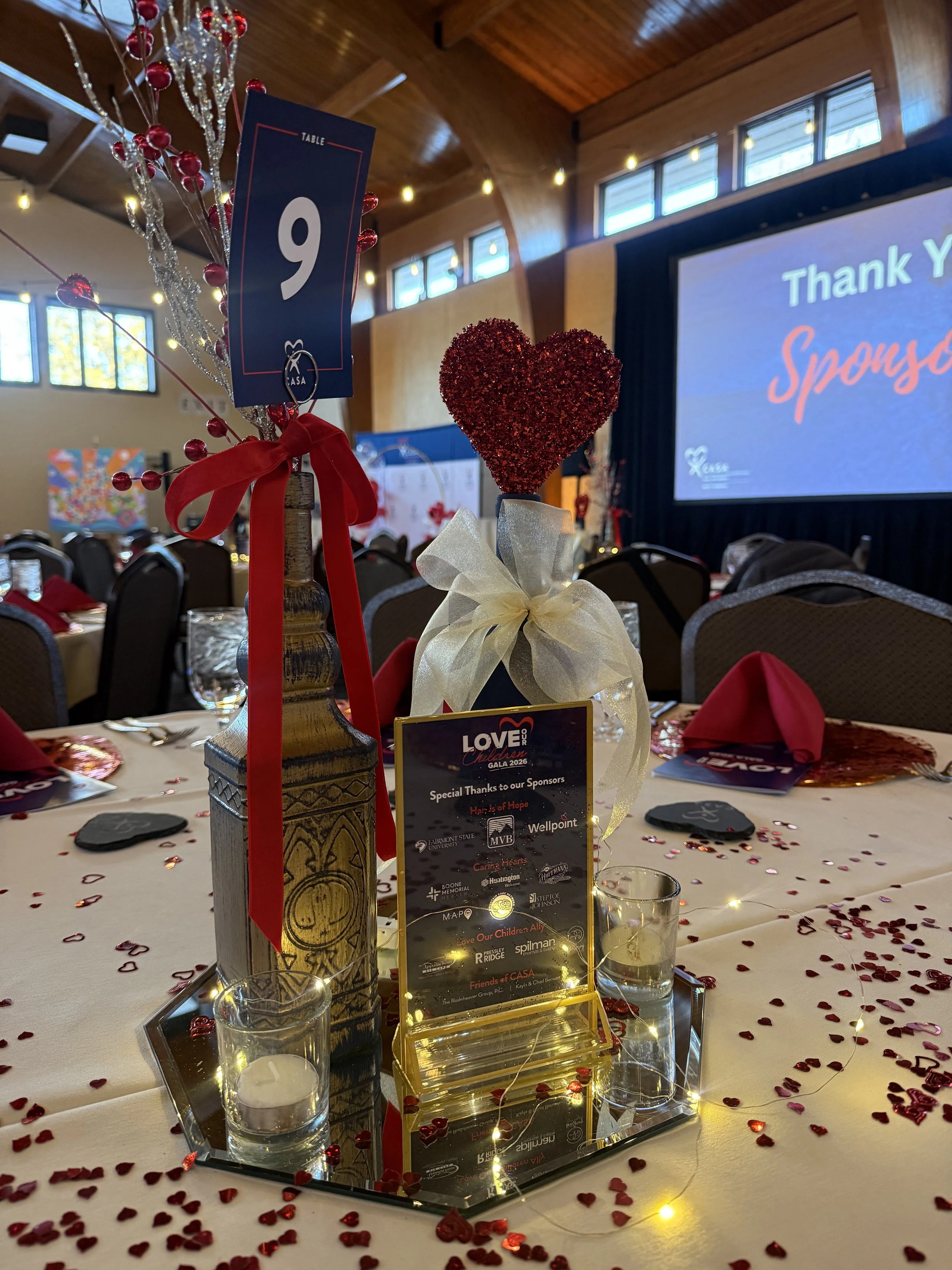 Table centerpiece with a red glitter heart decoration and a blue table number sign reading 9, surrounded by scattered red heart confetti, small candles, and an event program on a decorated banquet table inside a venue with wooden walls and large windows.