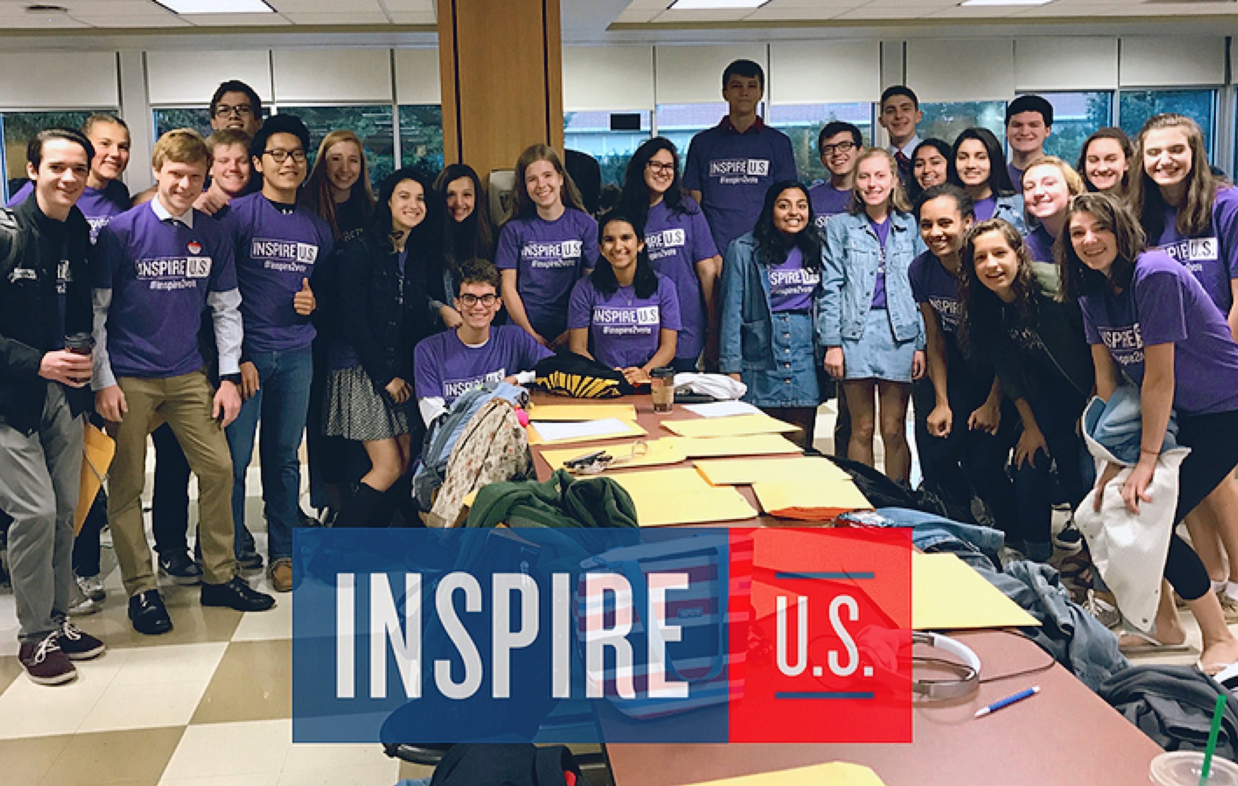 Group of young people at a meeting or event, some wearing purple shirts with 'INSPIRE U.S.' logos, standing in a room with tables and large windows.