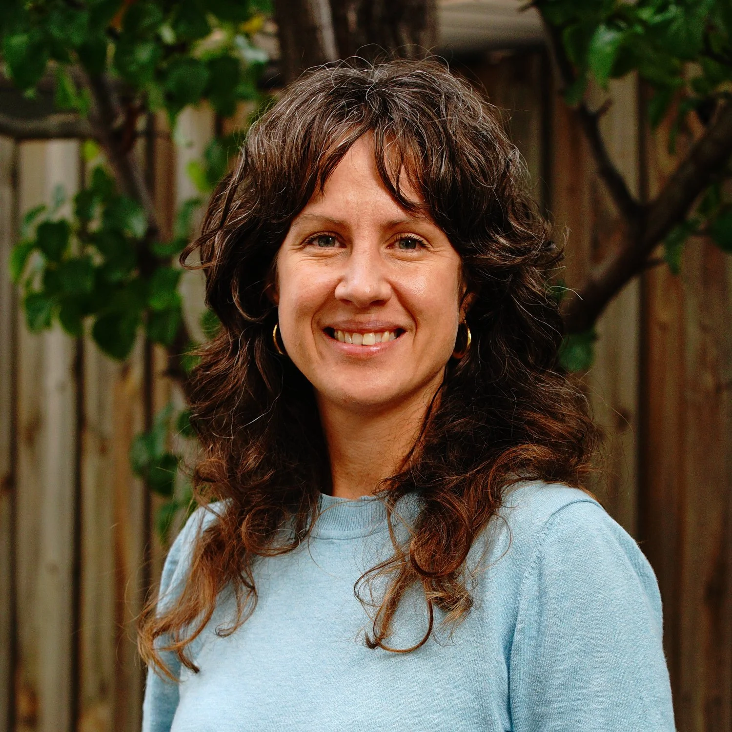 A woman with dark brown, curly hair smiling outdoors in front of a wooden fence and green foliage.