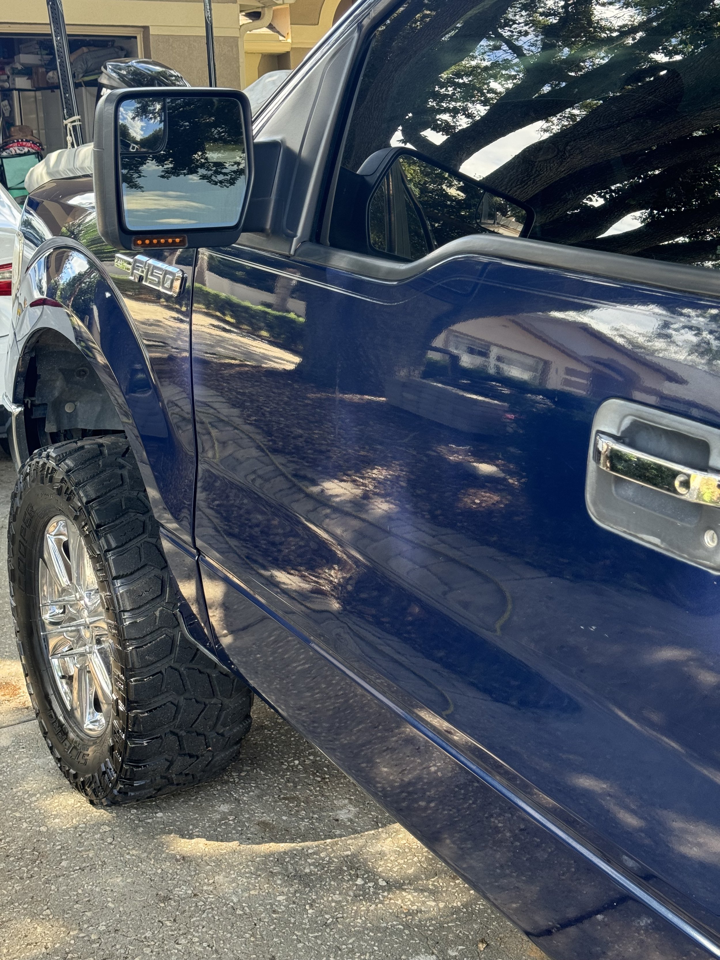 Close-up of the driver's side of a dark blue pickup truck, showing the side mirror, front door, and large off-road tire with chrome rim. Reflection of trees and a house can be seen on the truck's surface.