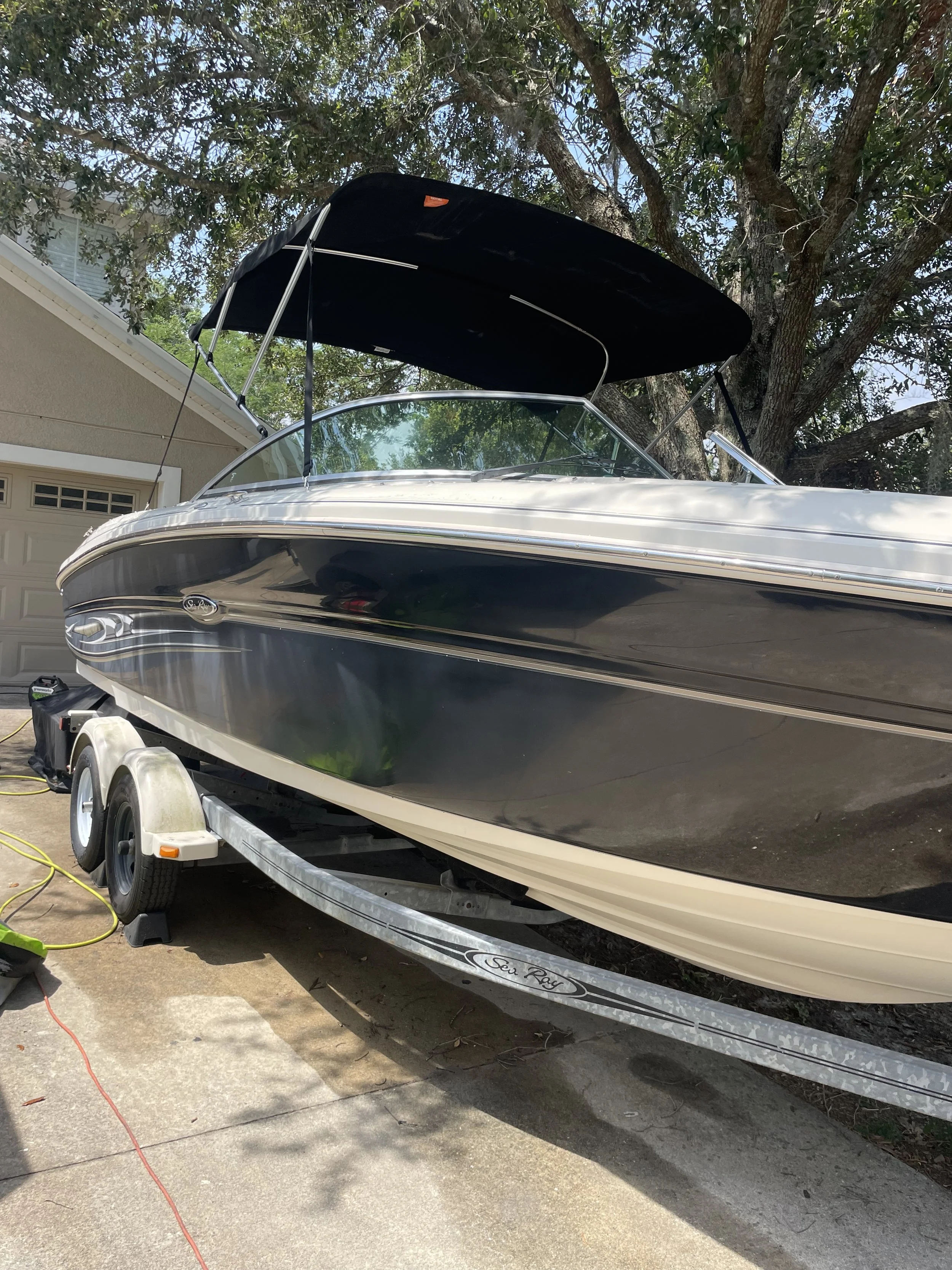 A boat on a trailer parked in a driveway, with a large tree and house in the background. The boat has a black and white exterior with a black canopy on top.
