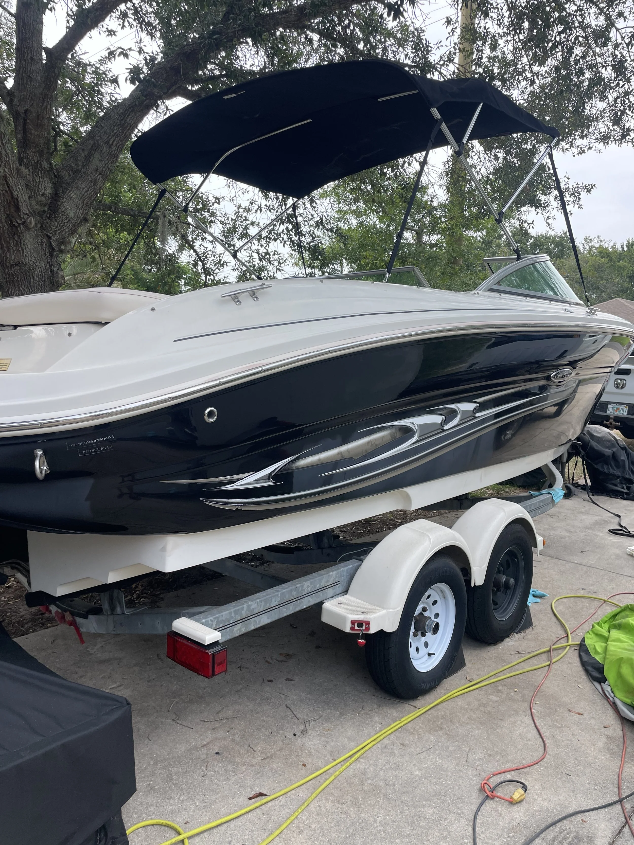 A black and white speedboat on a small trailer parked on a driveway surrounded by trees, with electrical cords and equipment nearby.