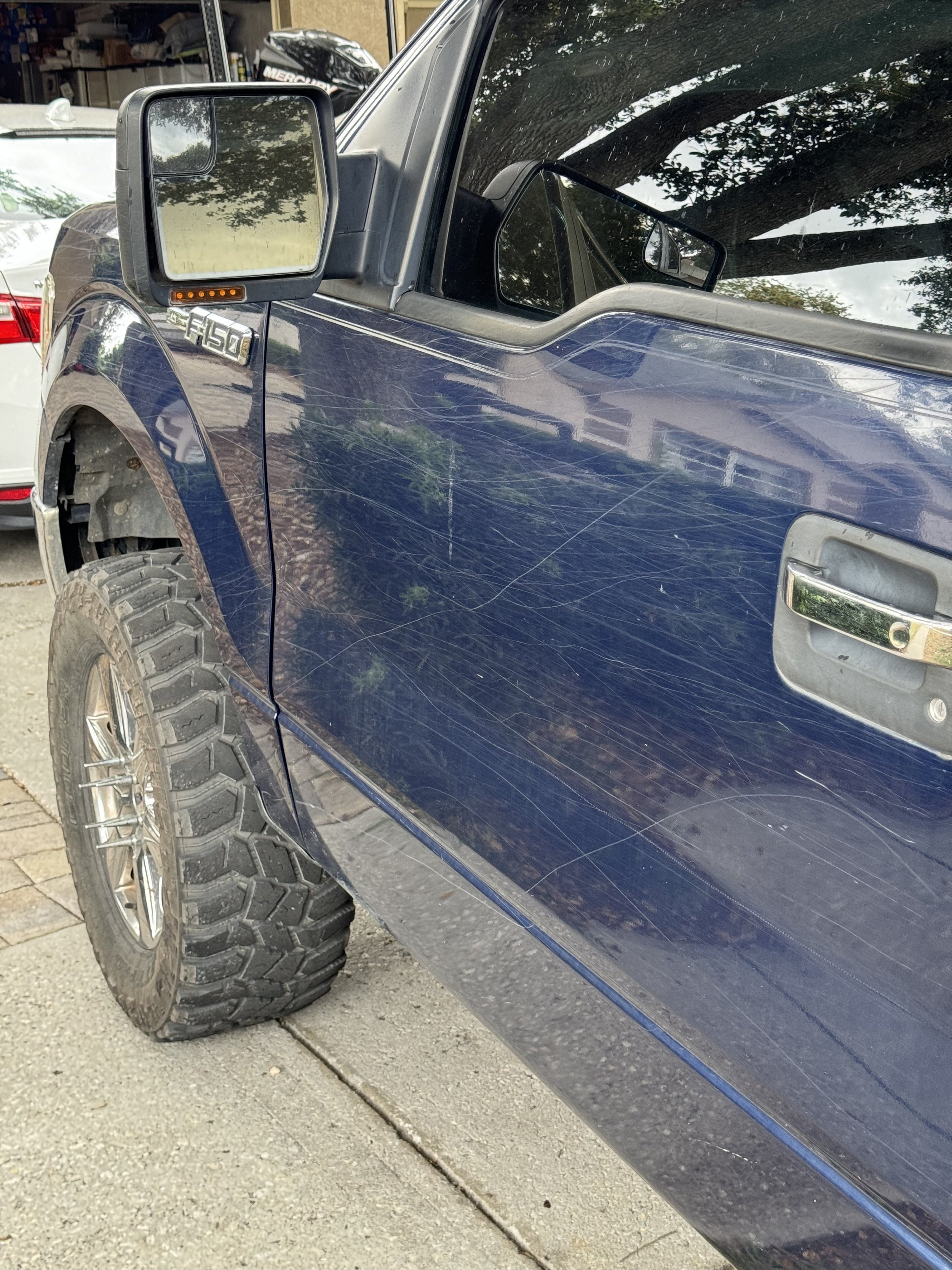 A blue pickup truck with extensive scratches on the front passenger side door, parked on a driveway. The driver’s side mirror is large and rectangular, showing reflections of trees and the sky. The front left tire is large with an off-road tread patt