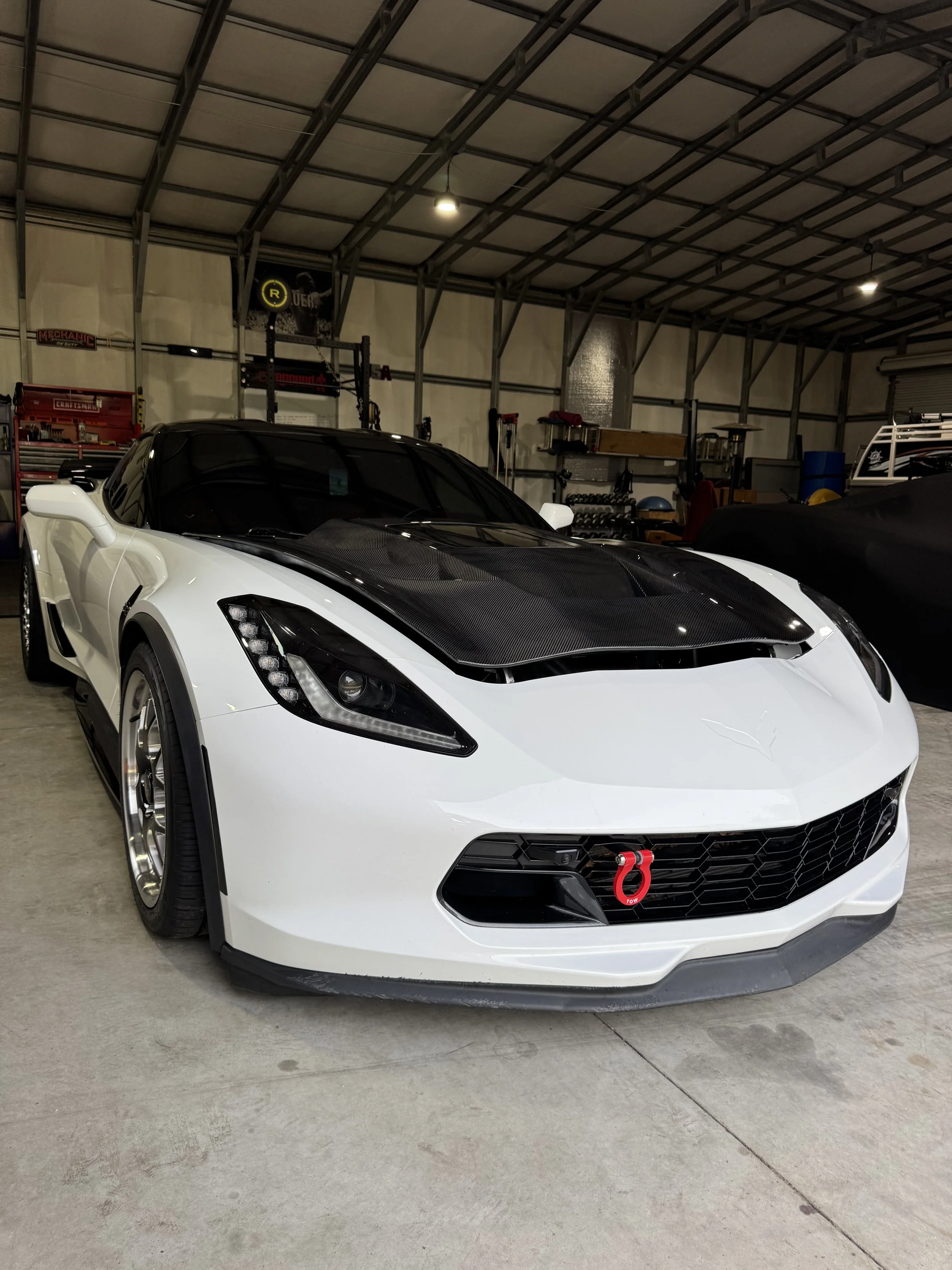 A white sports car with a black carbon fiber hood, parked inside a garage. The car has a red tow hook at the front. The garage has various tools and equipment along the back wall, with a metal shelving unit and a workbench.