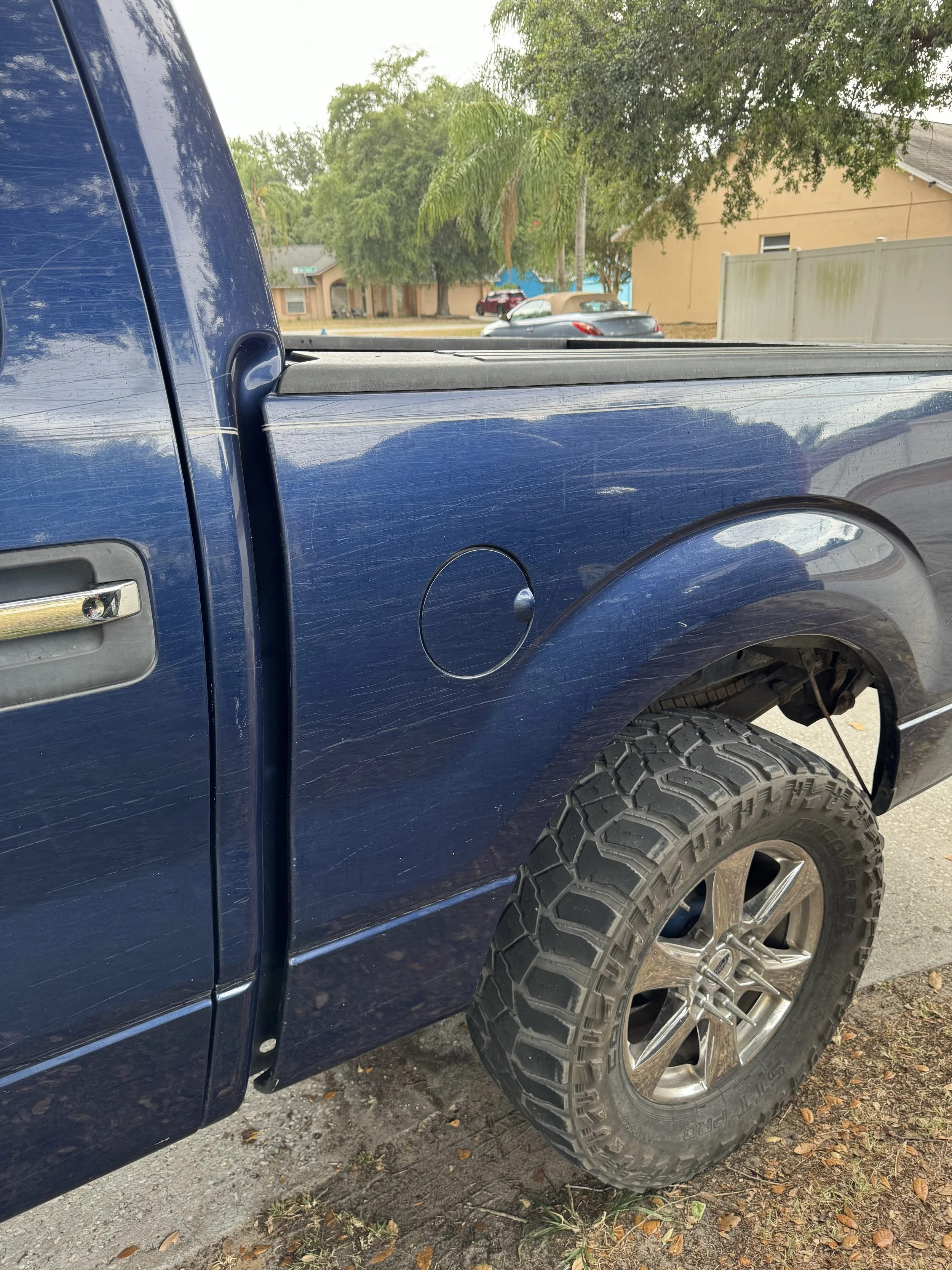 Close-up of a blue truck with a scratched door, large off-road tire, and part of the truck bed in an outdoor residential area with leaves on the ground, trees, and houses in the background.