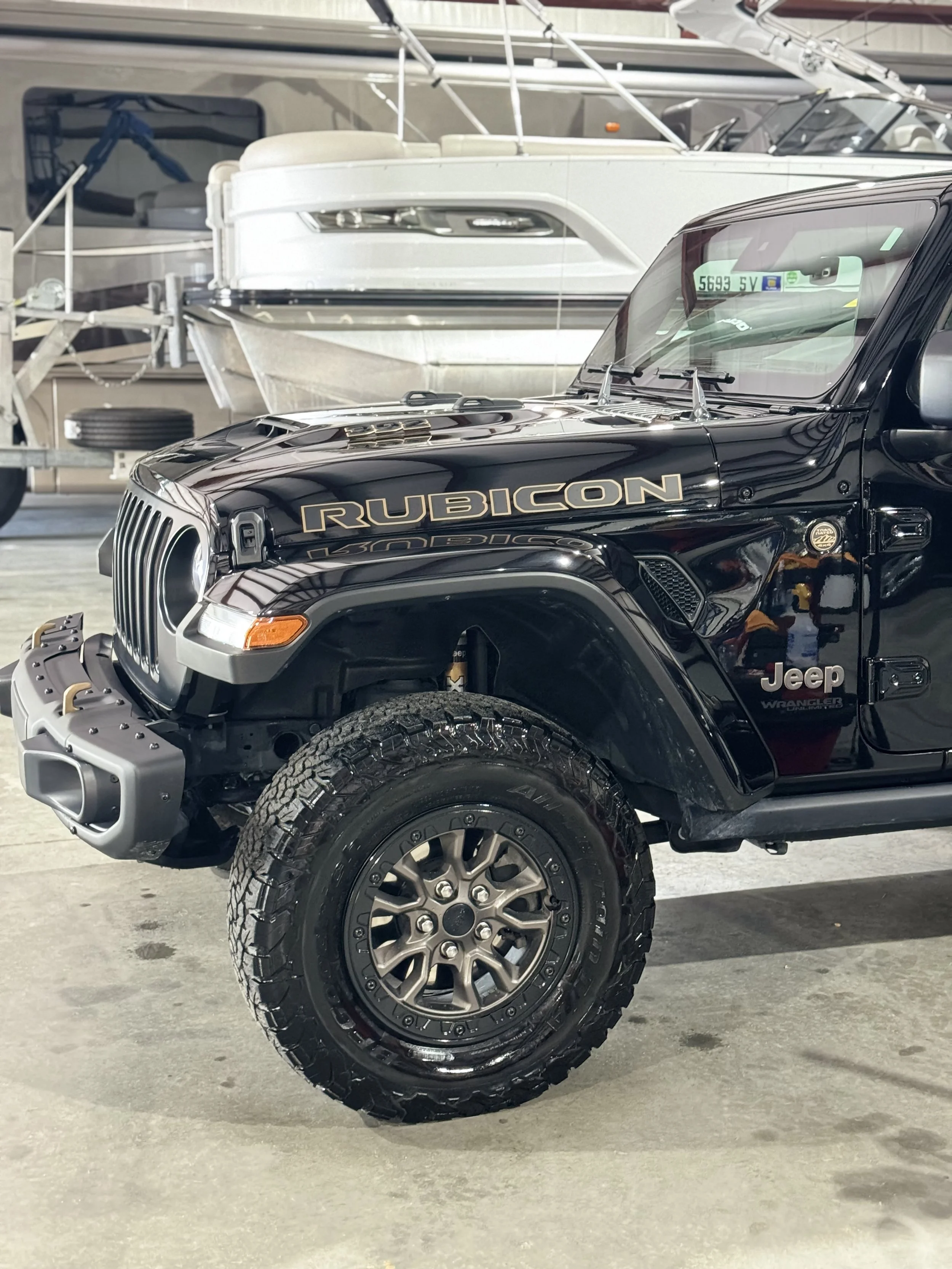 Close-up of a black Jeep Rubicon Wrangler with a prominent gold 'RUBICON' logo on the hood, showing rugged off-road tires and chrome accents, parked in an indoor garage with a boat in the background.