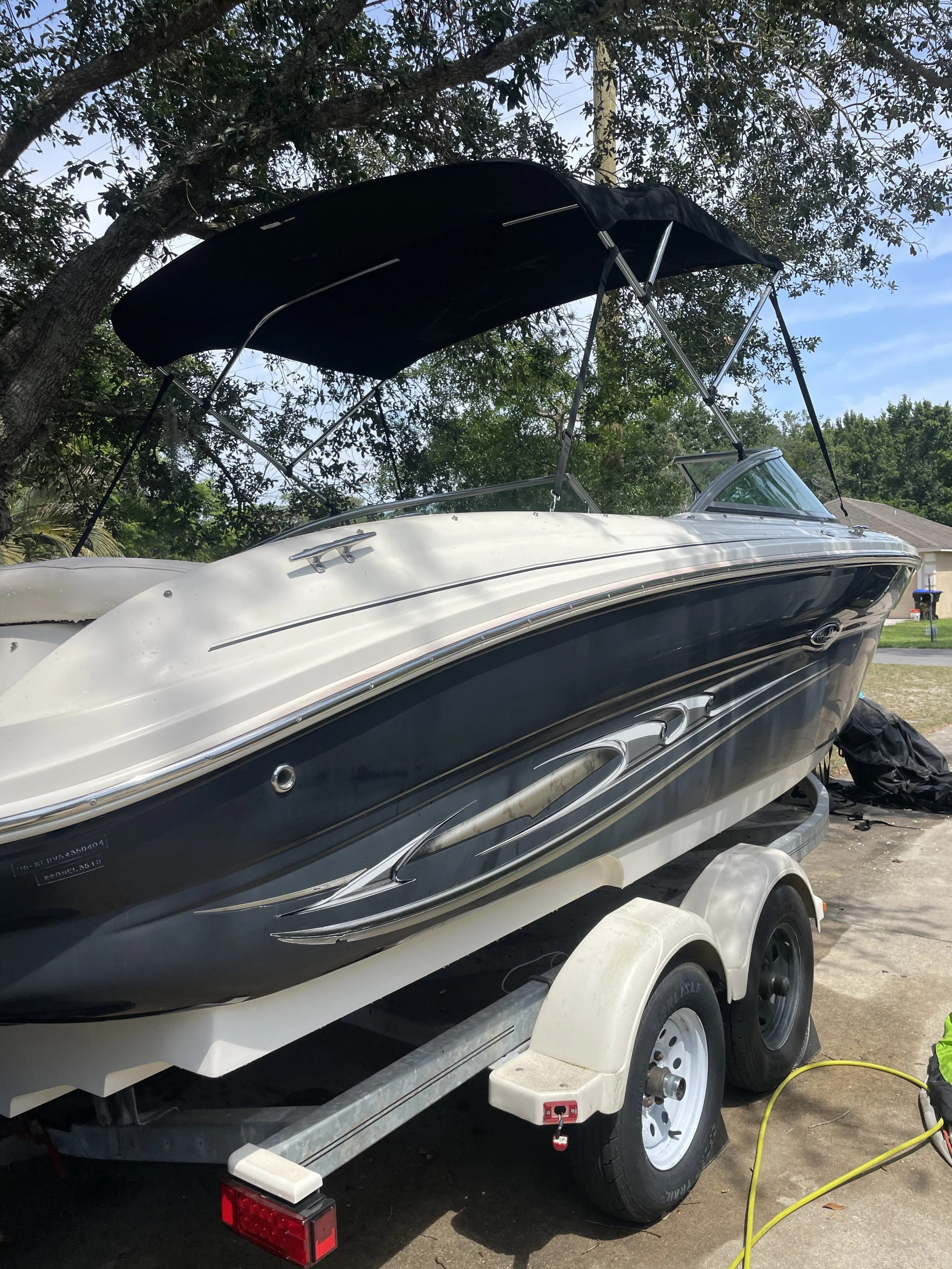 A motorboat on a trailer, parked on a driveway, with a black canopy and a white hull, connected to an electric cord, near trees and a house in the background.