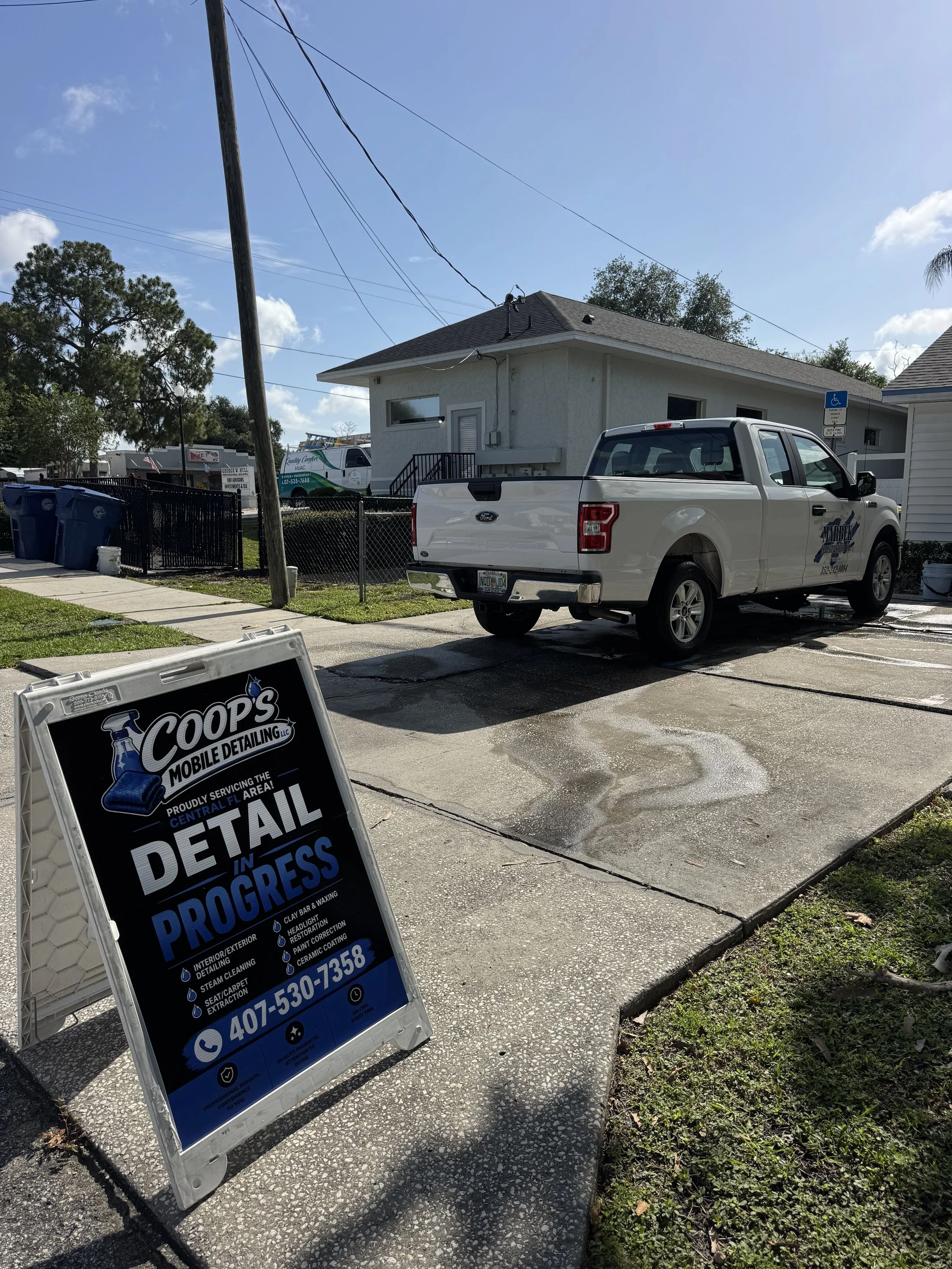 Sign for progress in detail car wash and detailing at a sidewalk in front of a business, with a white pickup truck parked nearby, a house in the background, blue trash bins, and utility poles with power lines.
