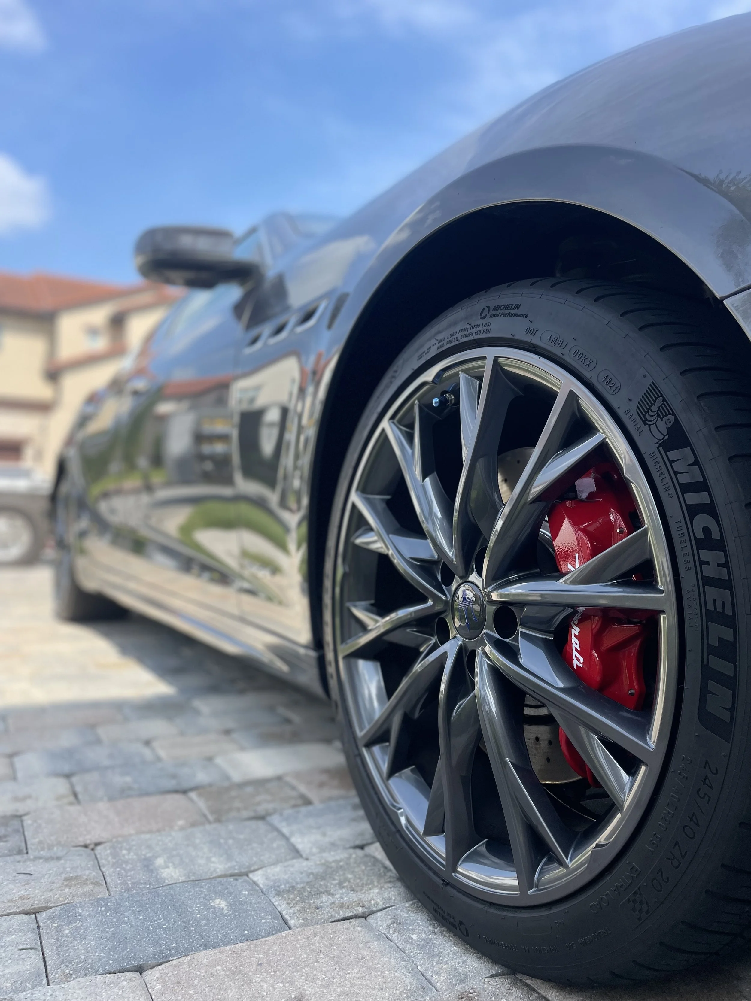 Close-up of a sleek black sports car wheel with a red brake caliper, parked on a driveway with cobblestone pavement, houses and a blue sky in the background.