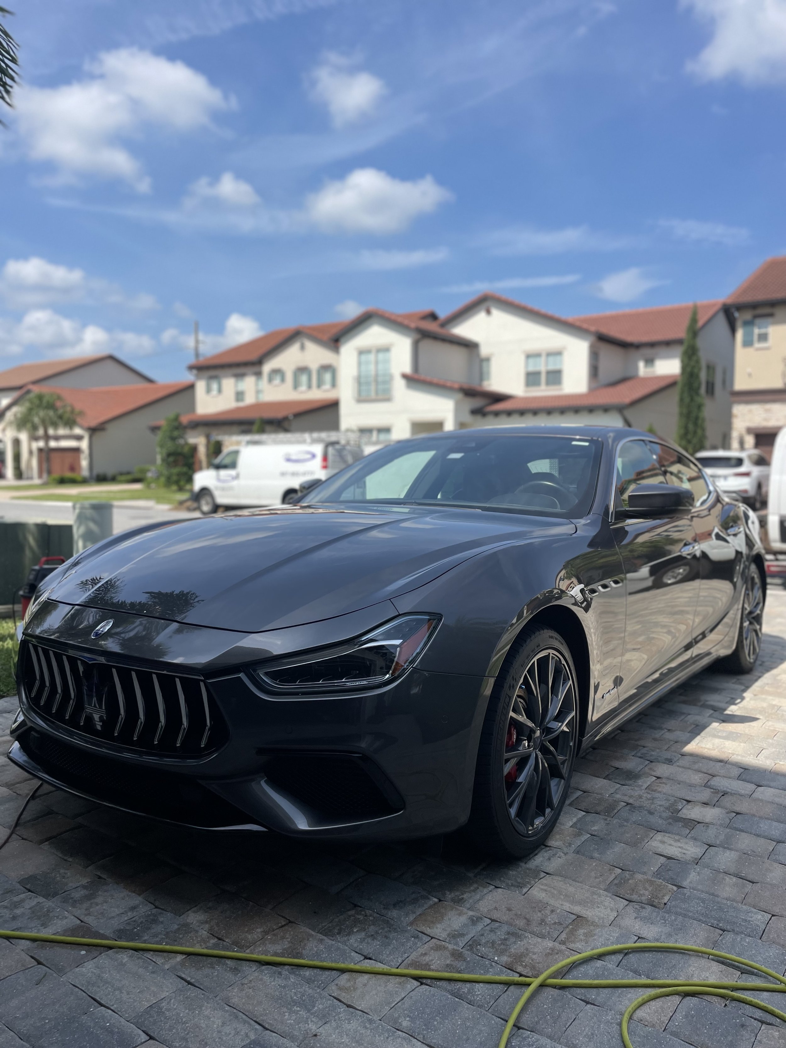 Black sports car parked on a stone driveway in front of suburban houses under a blue sky with clouds.