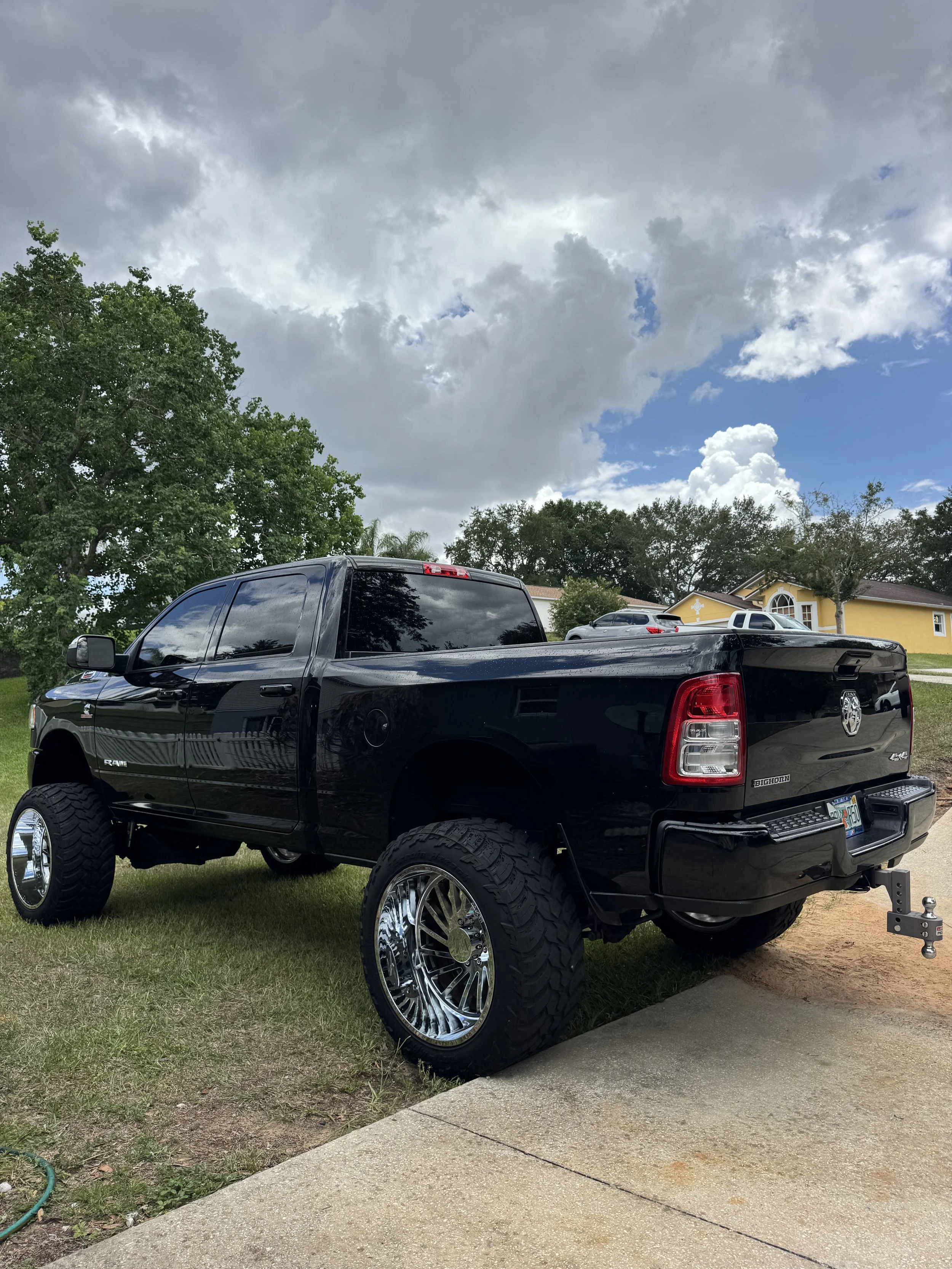 A black pickup truck with large chrome wheels parked on a suburban lawn. There are other vehicles in the background and trees along with a yellow house. The sky is partly cloudy with blue showing through.