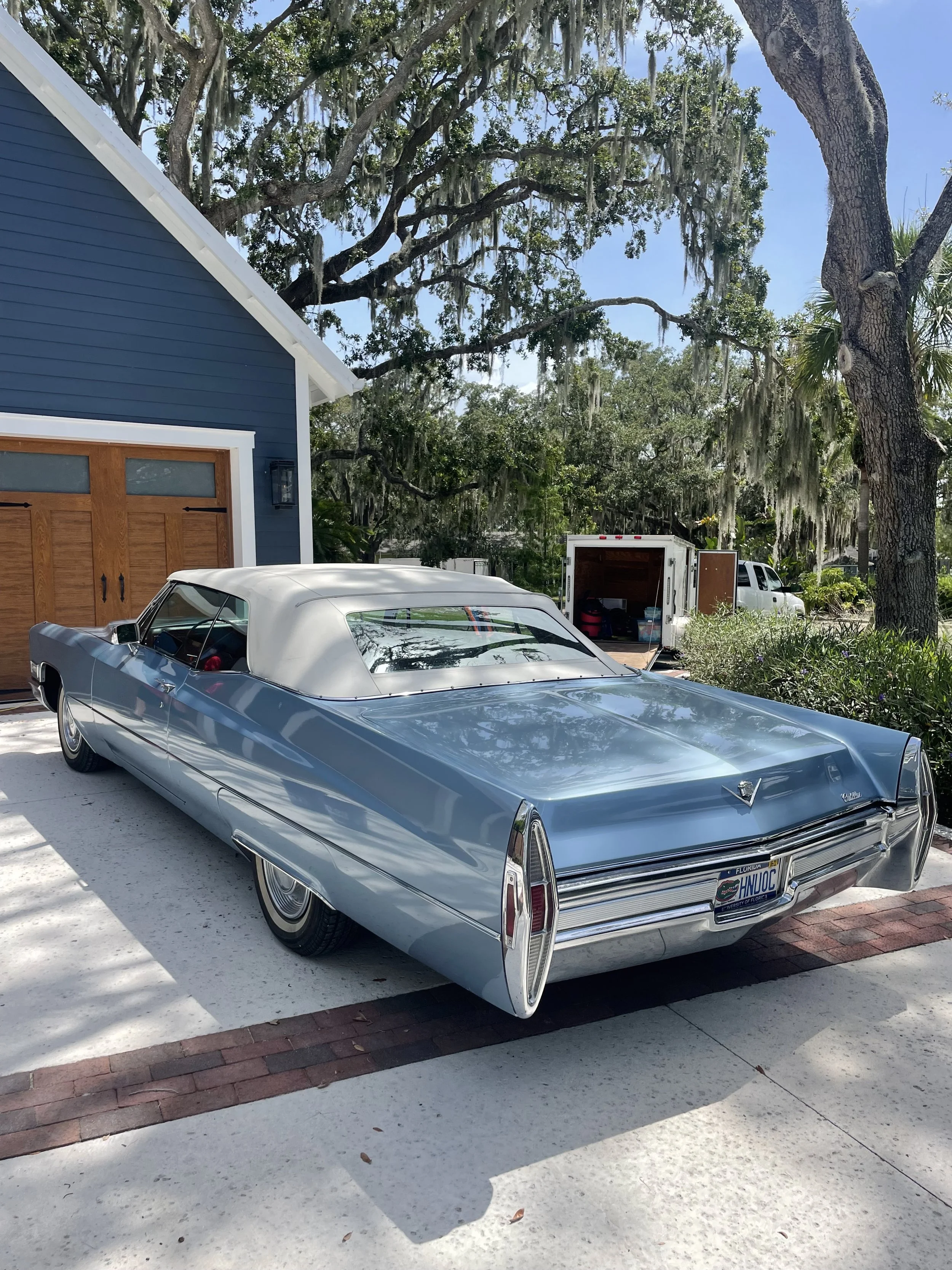 A vintage blue car with a white roof parked in a driveway in front of a blue house with wooden garage doors, surrounded by trees with Spanish moss and a white utility trailer.