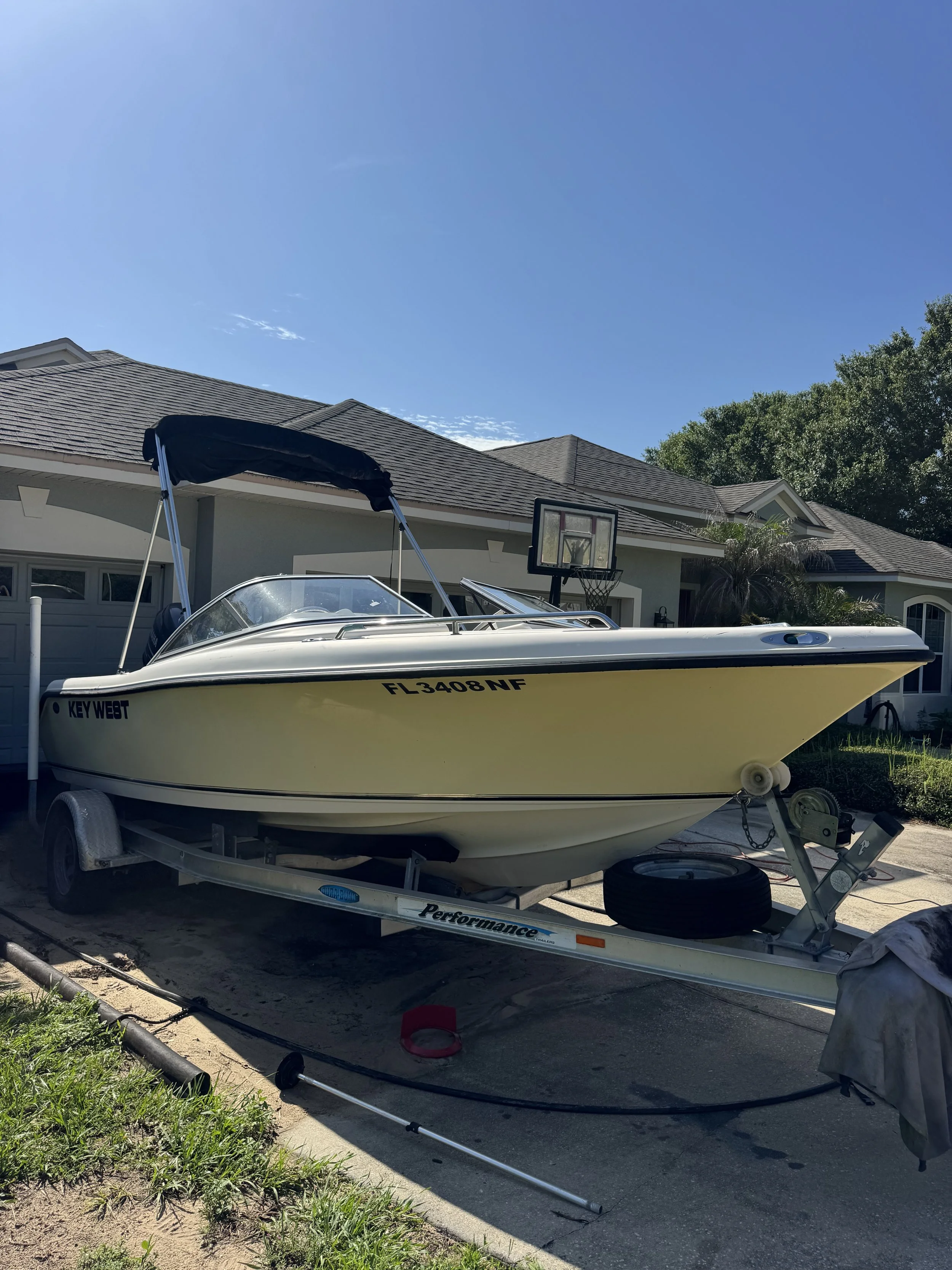 A boat on a trailer parked in a driveway in front of a house with a basketball hoop. The house has a light-colored exterior and a dark shingle roof. The boat is white with a black canopy and is facing towards the street. There are some gardening tool