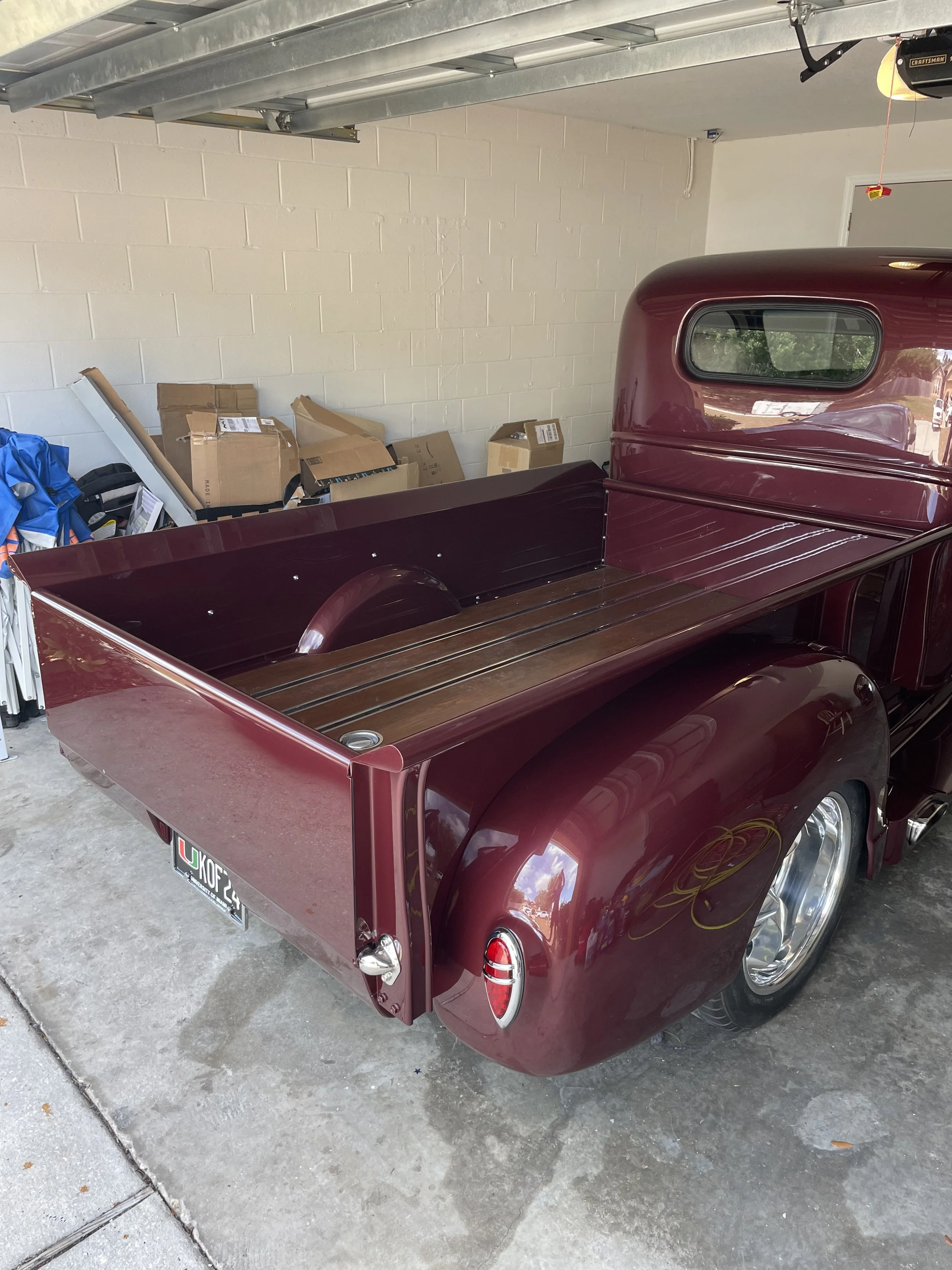 A vintage maroon pickup truck parked in a garage with a white brick wall, cardboard boxes, and miscellaneous items stacked against the wall.