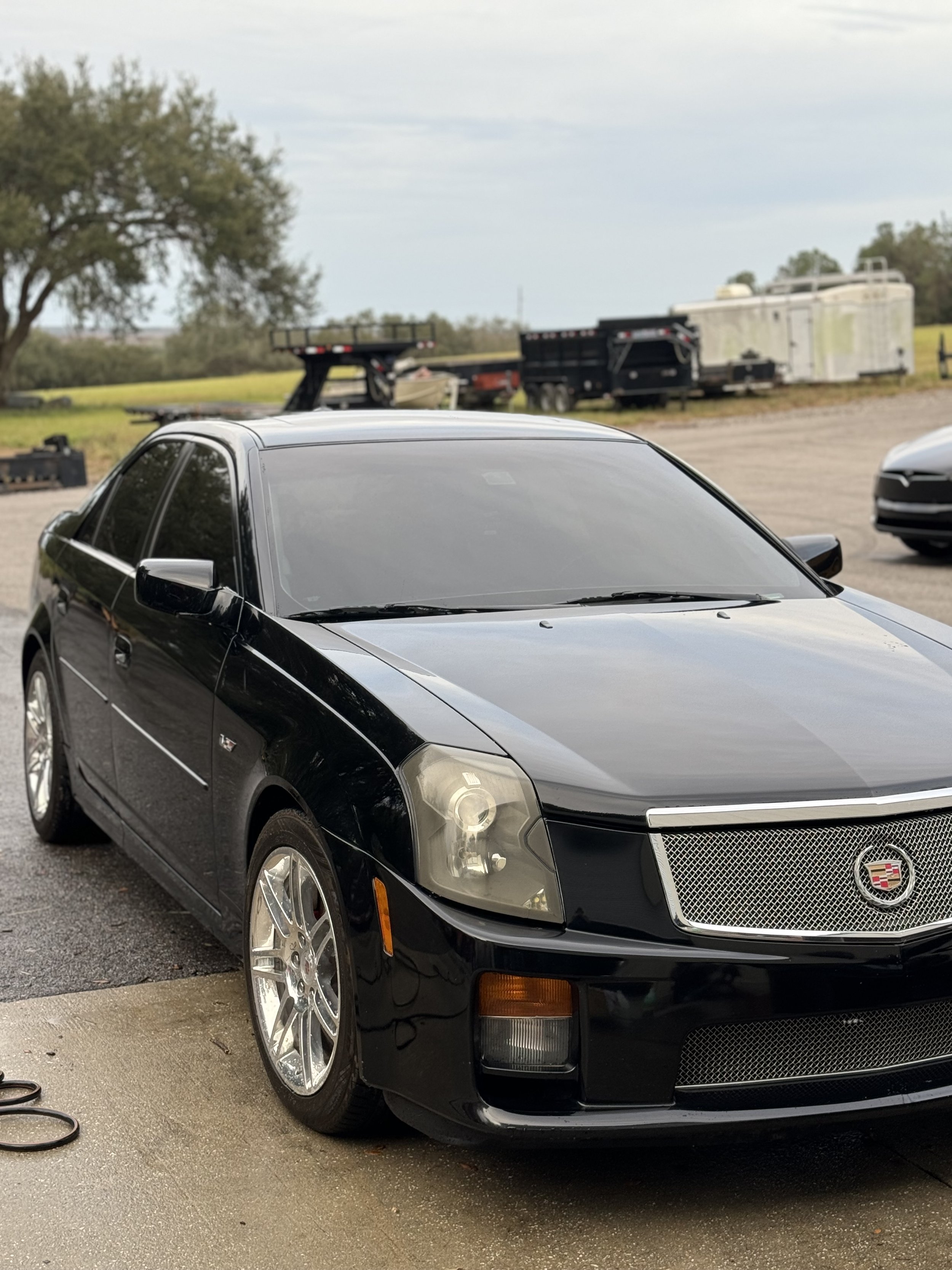 Black Cadillac car parked in a driveway with a trailer and trees in the background.