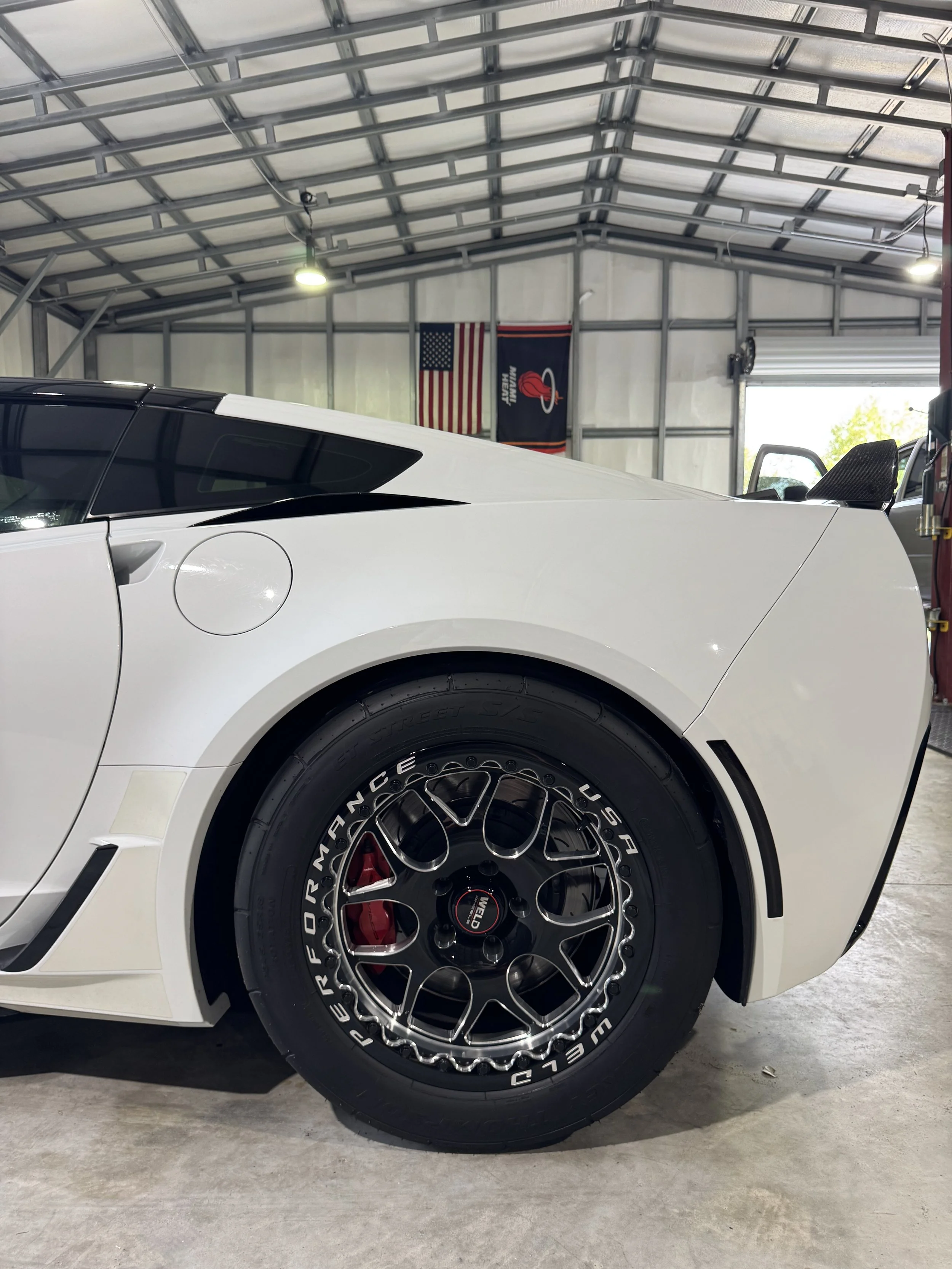 Close-up of a white sports car inside a garage, showing the rear wheel with black, multi-spoke rim and tire branded 'WELD Racing'. The garage has metal framing, American flag, and a flag with Rocket League logo on the wall.