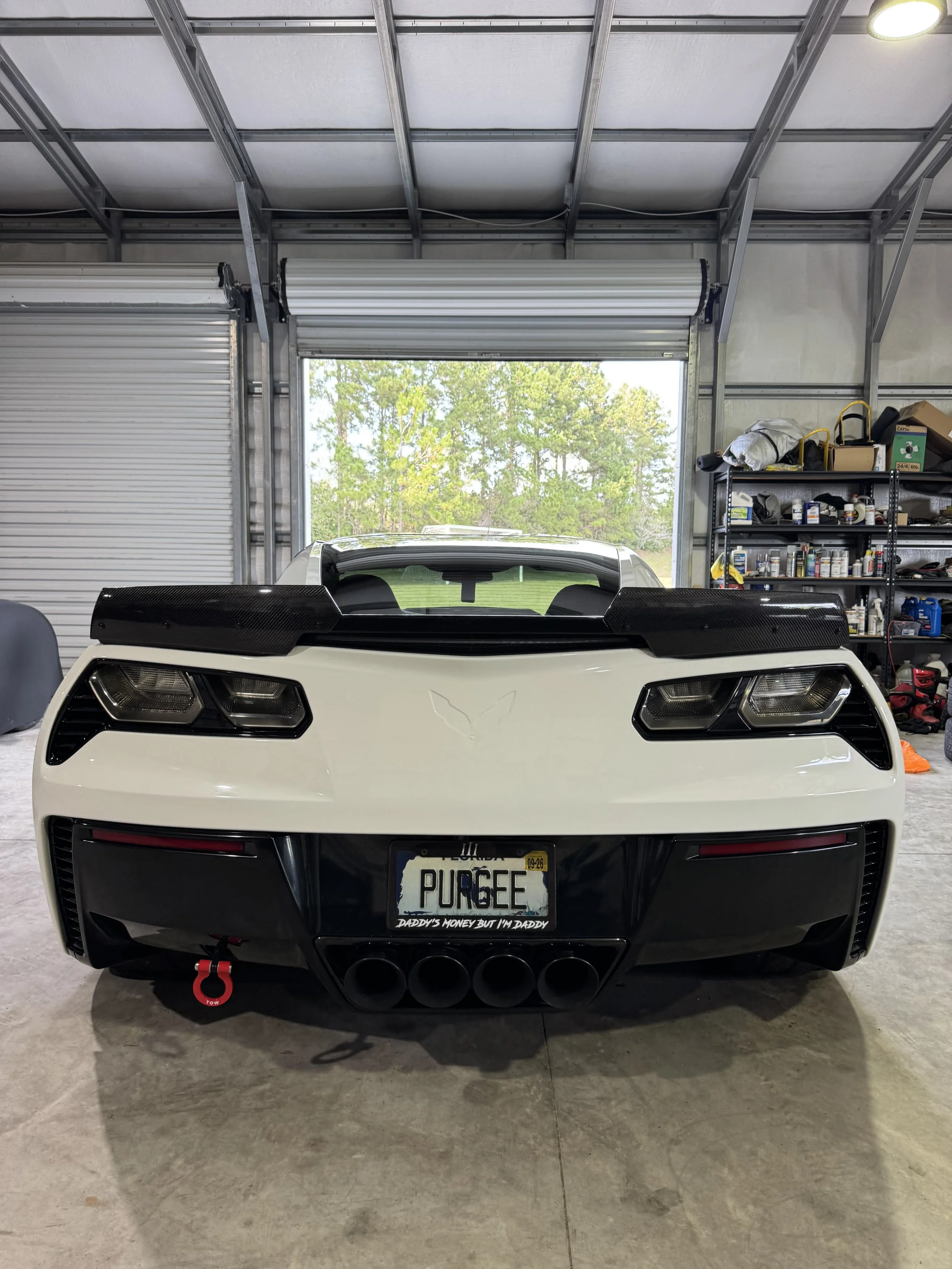 Front view of a white sports car inside a garage, with a custom license plate reading 'PURGE' and a bumper sticker that says 'Daddy's money but I'm daddy', showing a rear spoiler, black exhaust pipes, and a red tow hook, with an open garage door in t
