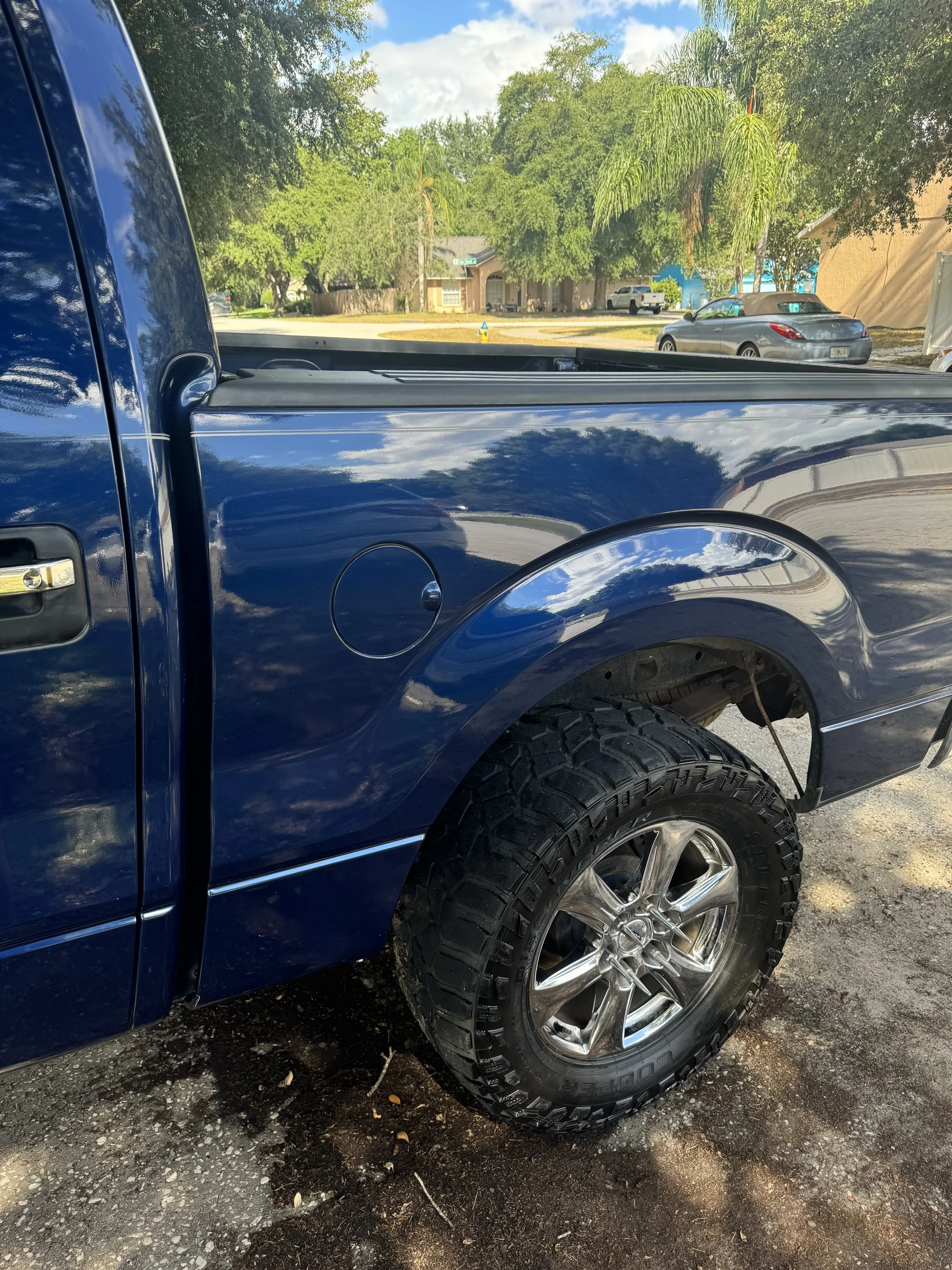 Close-up of a dark blue pickup truck with a lifted tire and chrome rim, parked on a driveway with trees and a house in the background.